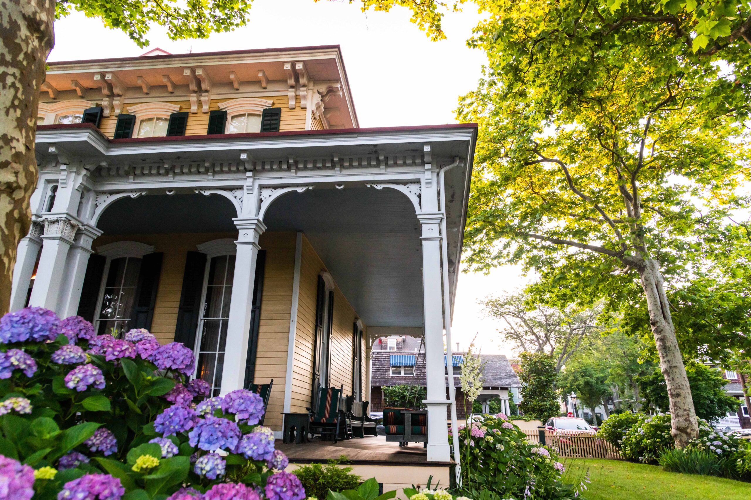 Sunlight shining behind trees and purple hydrangea around the Mainstay Inn, a yellow Victorian styled bed and breakfast.