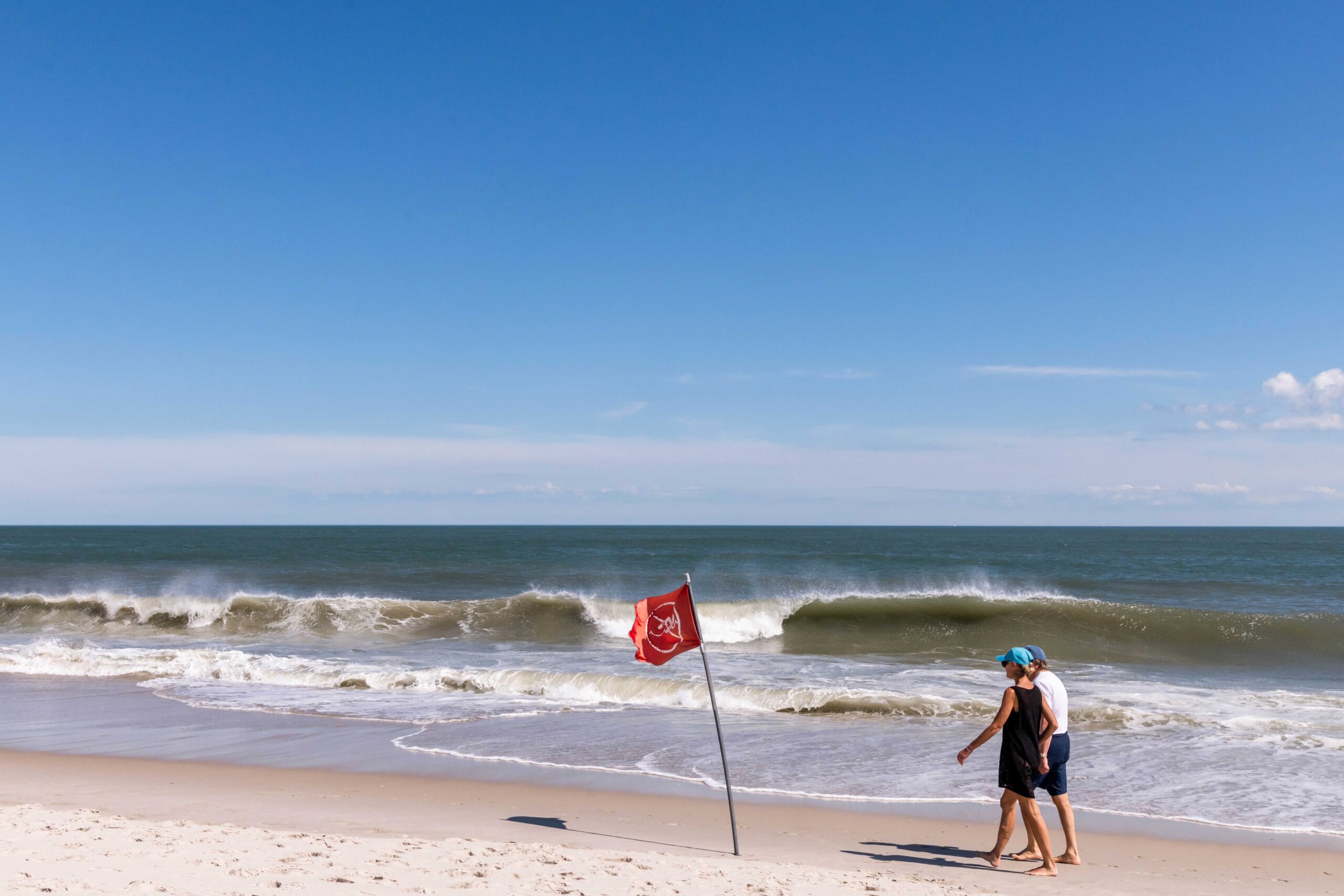 Two people walking on the beach on a sunny day with a bright blue sky. A wave is crashing in the ocean and a red don't swim flag on the beach is blowing in the breeze.