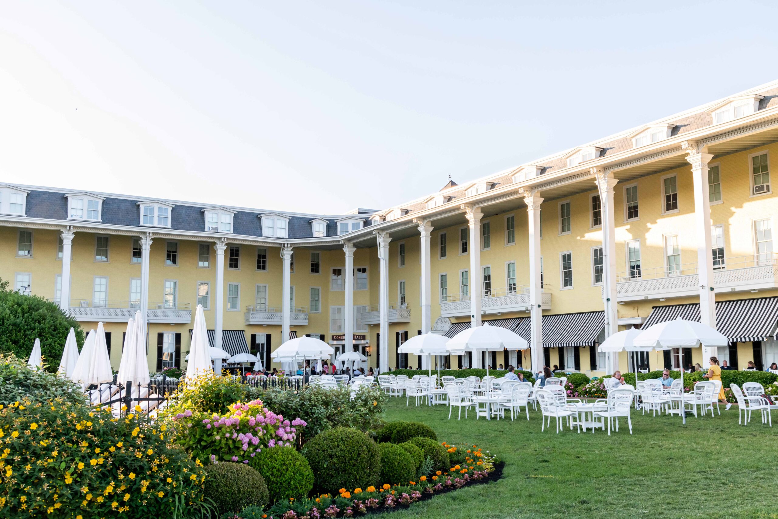 The front lawn of Congress Hall (a yellow L shaped Victorian hotel with white pillars) with white tables, chairs and umbrellas and flowers on a sunny day
