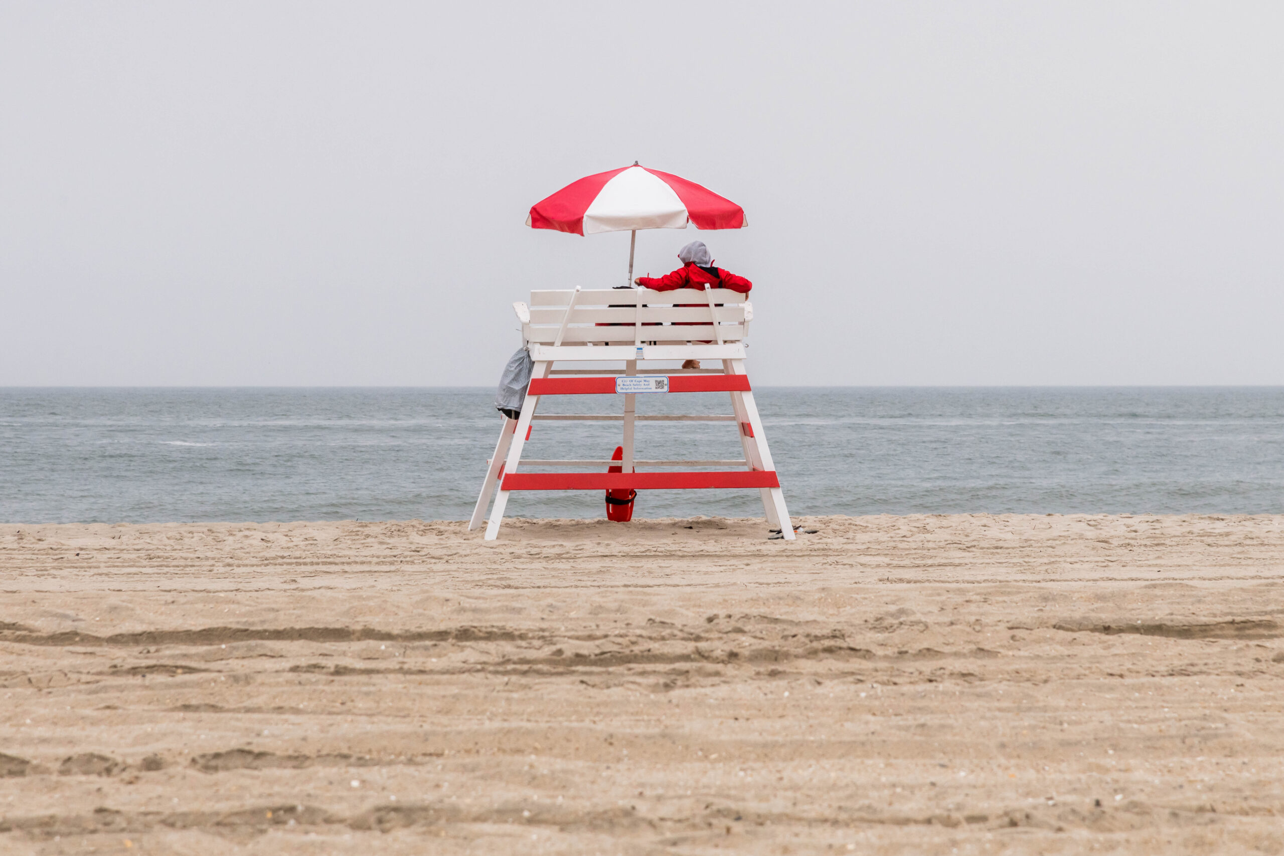 A lifeguard wearing a red jacket sitting on a red and white lifeguard stand under a red and white umbrella on the beach with a cloudy gray sky and gray sea