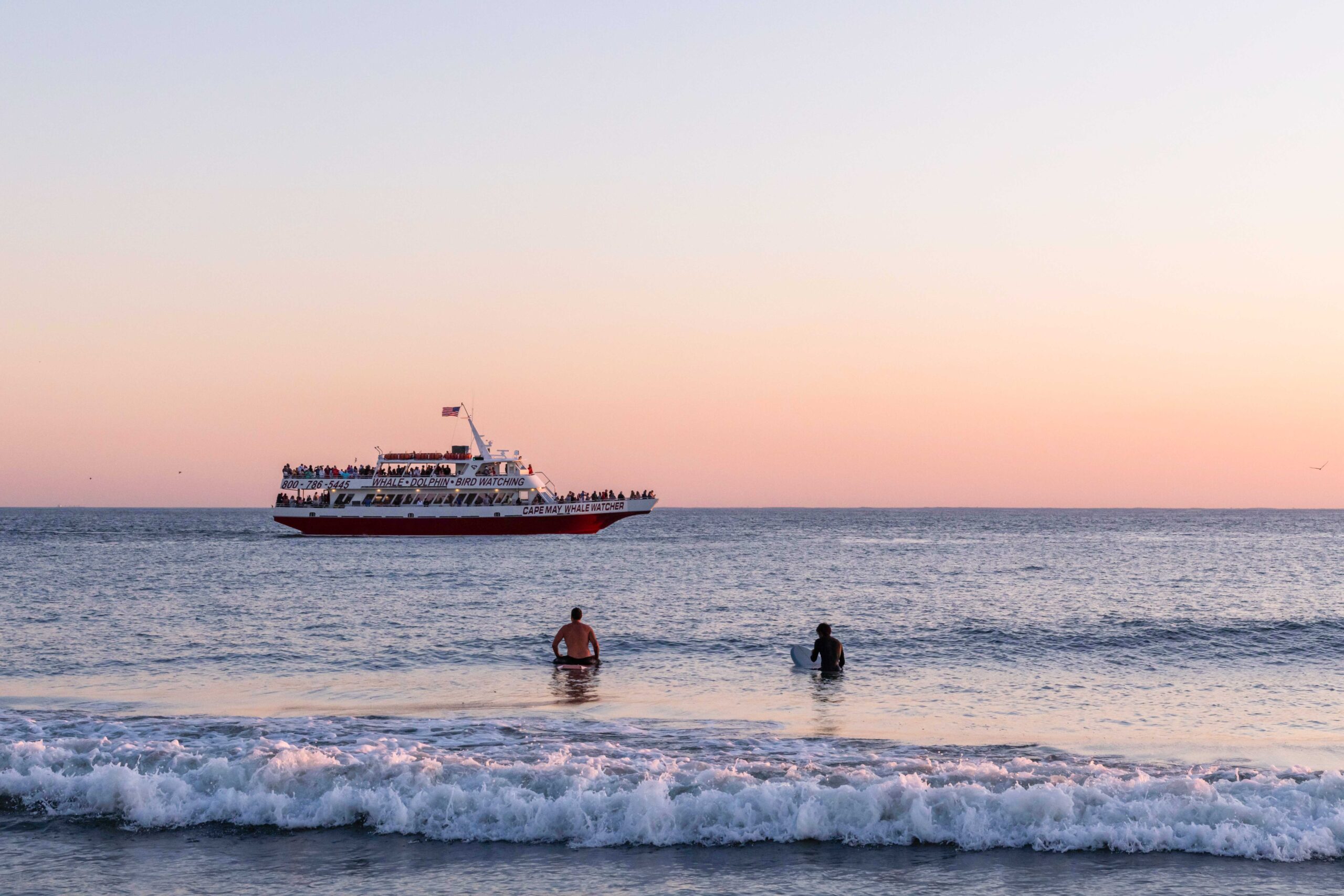Two people sitting on surfboards in the ocean watching the Cape May Whale Watching boat sail past at sunset