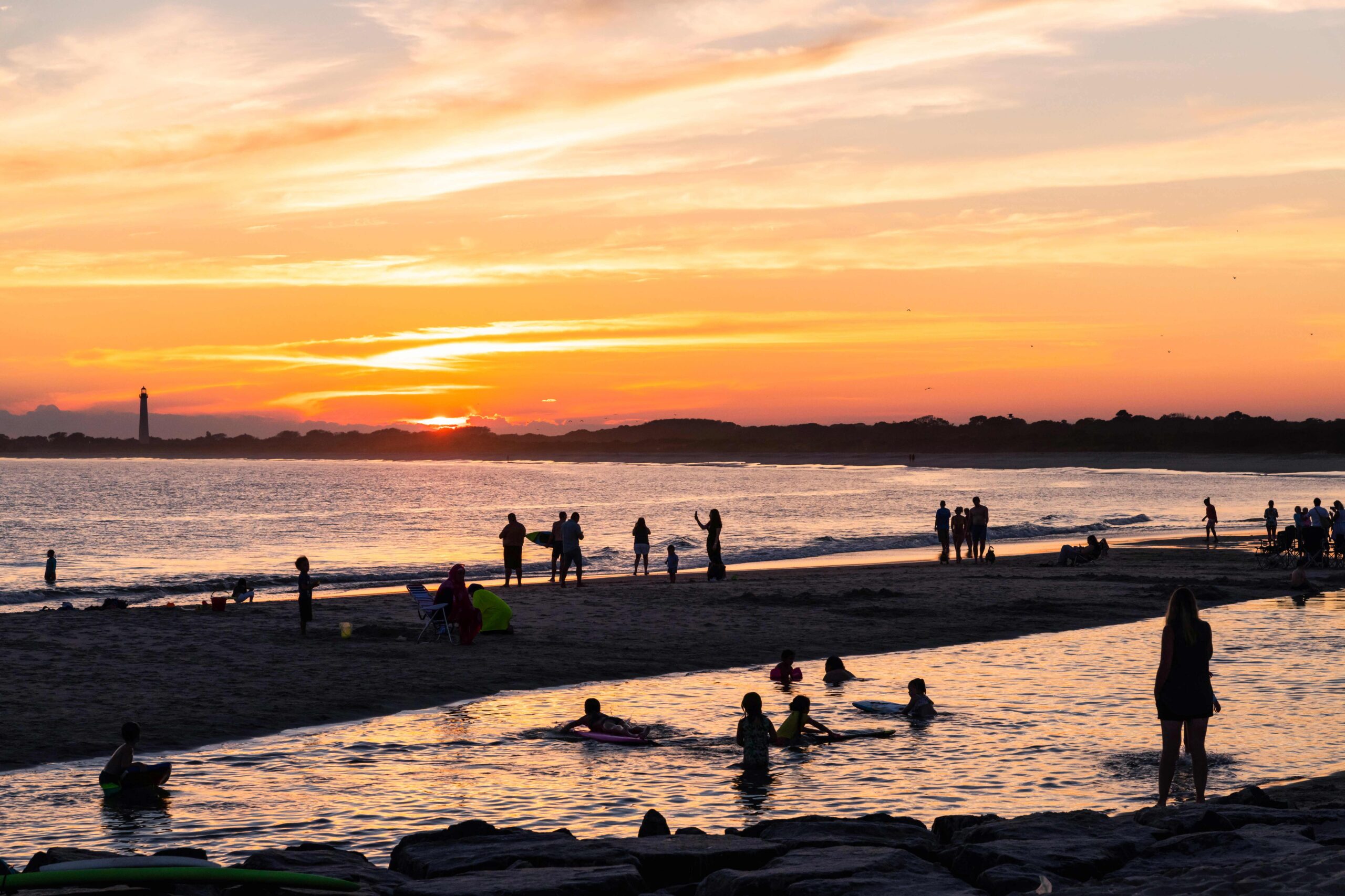 People swimming and watching sunset at the beach with the sun setting and the Cape May Lighthouse in the distance