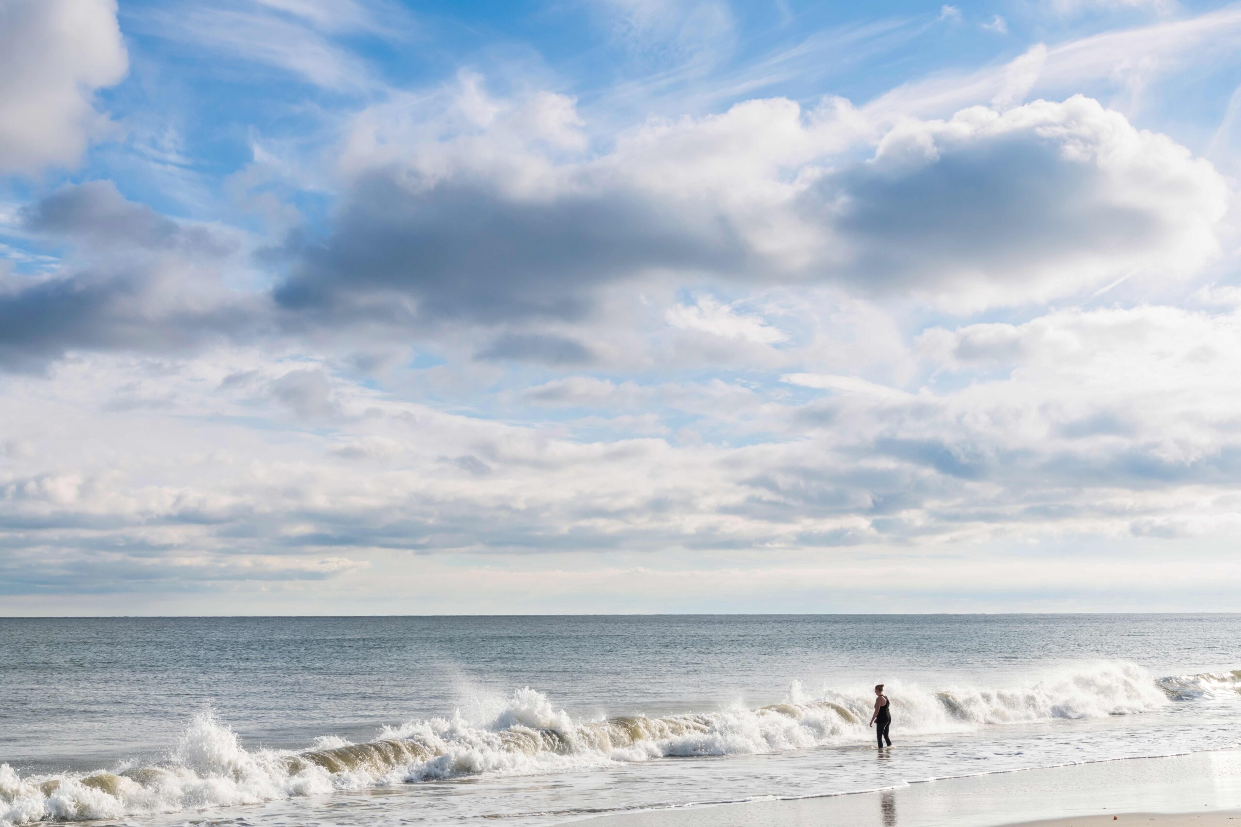 A person going into the ocean in the morning with a wave crashing and some puffy clouds in the sky