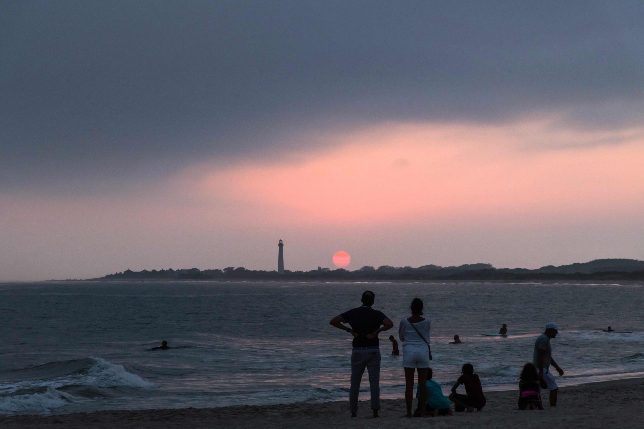 People on the beach and swimming in the ocean watching a pink sunset with a dark cloud in the sky