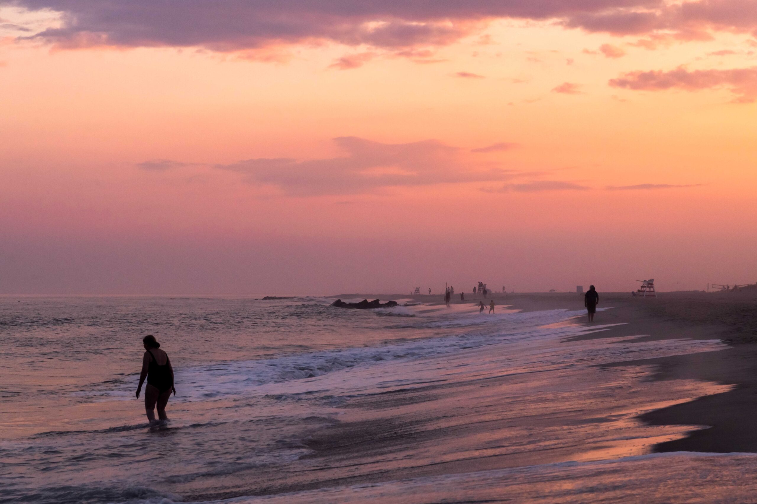 A person walking into the ocean and other people in the distance with a pink sky and some wispy purple and pink clouds