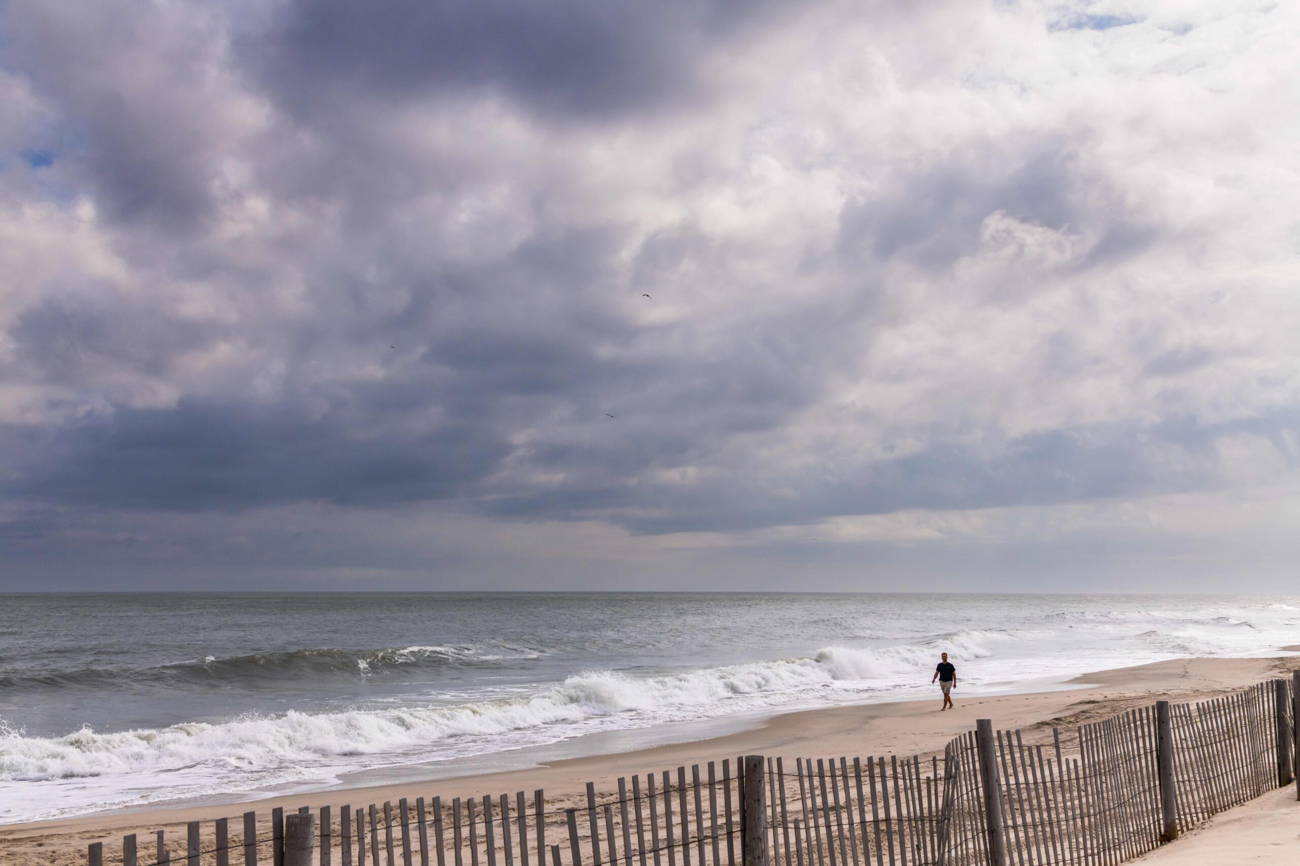 A person walking on the beach with clouds on the sky, a wave crashing, and the beach fence in the foreground