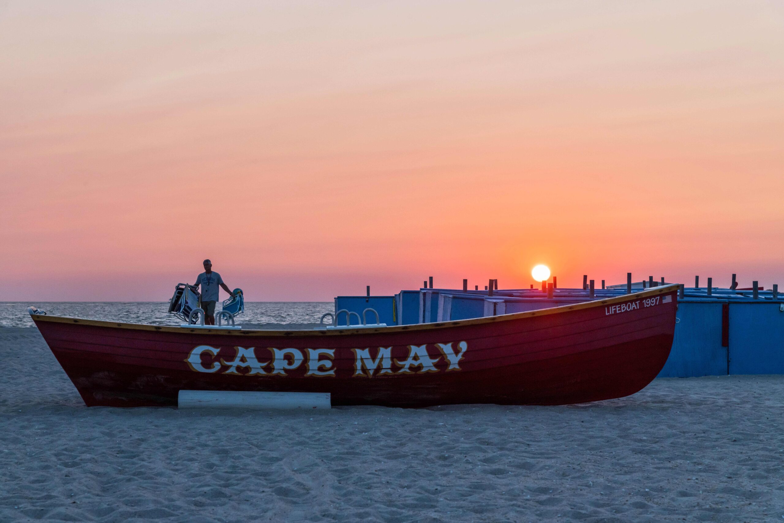 The sun setting behind blue Steger's beach boxes and a red Cape May lifeguard boat with a person carrying beach chairs in the background