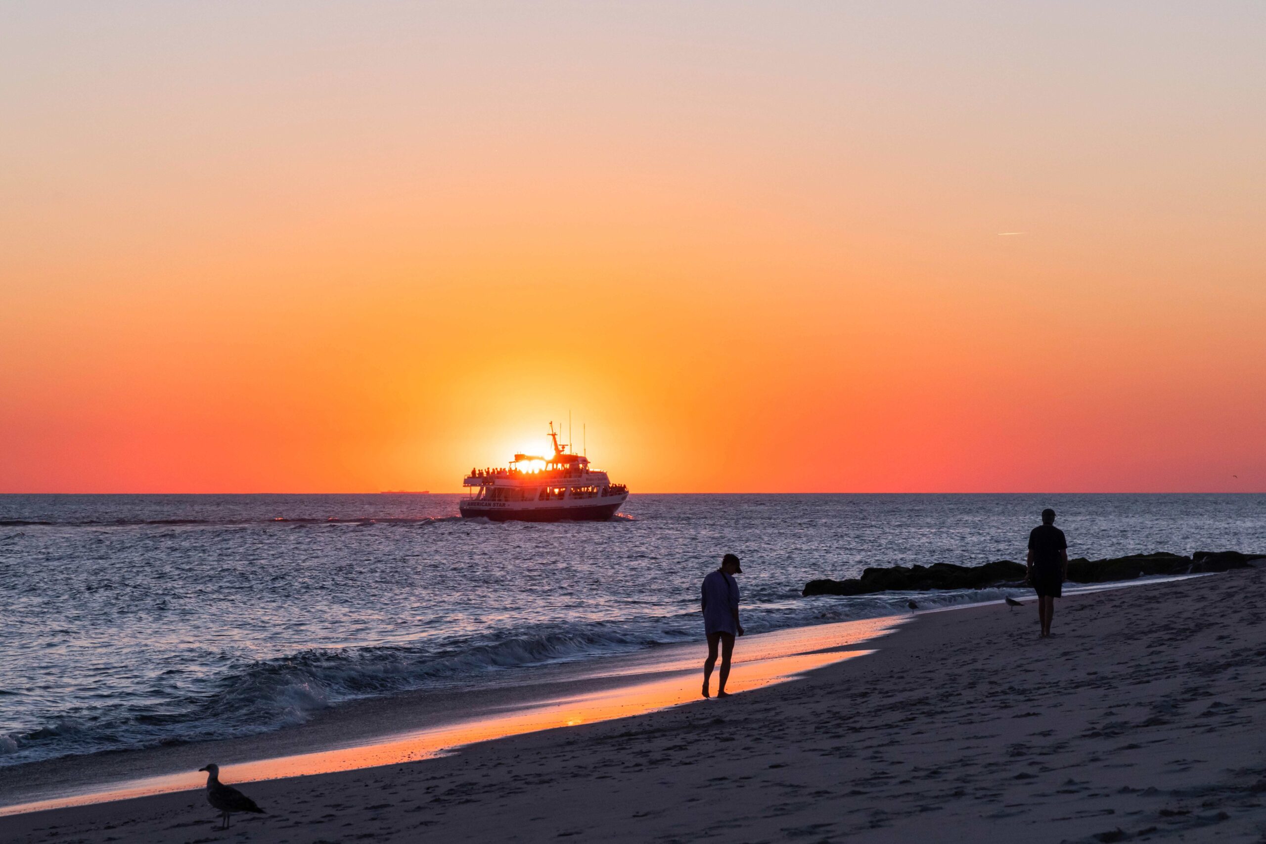 Two people walking on the beach with a boat sailing past the sun at sunset