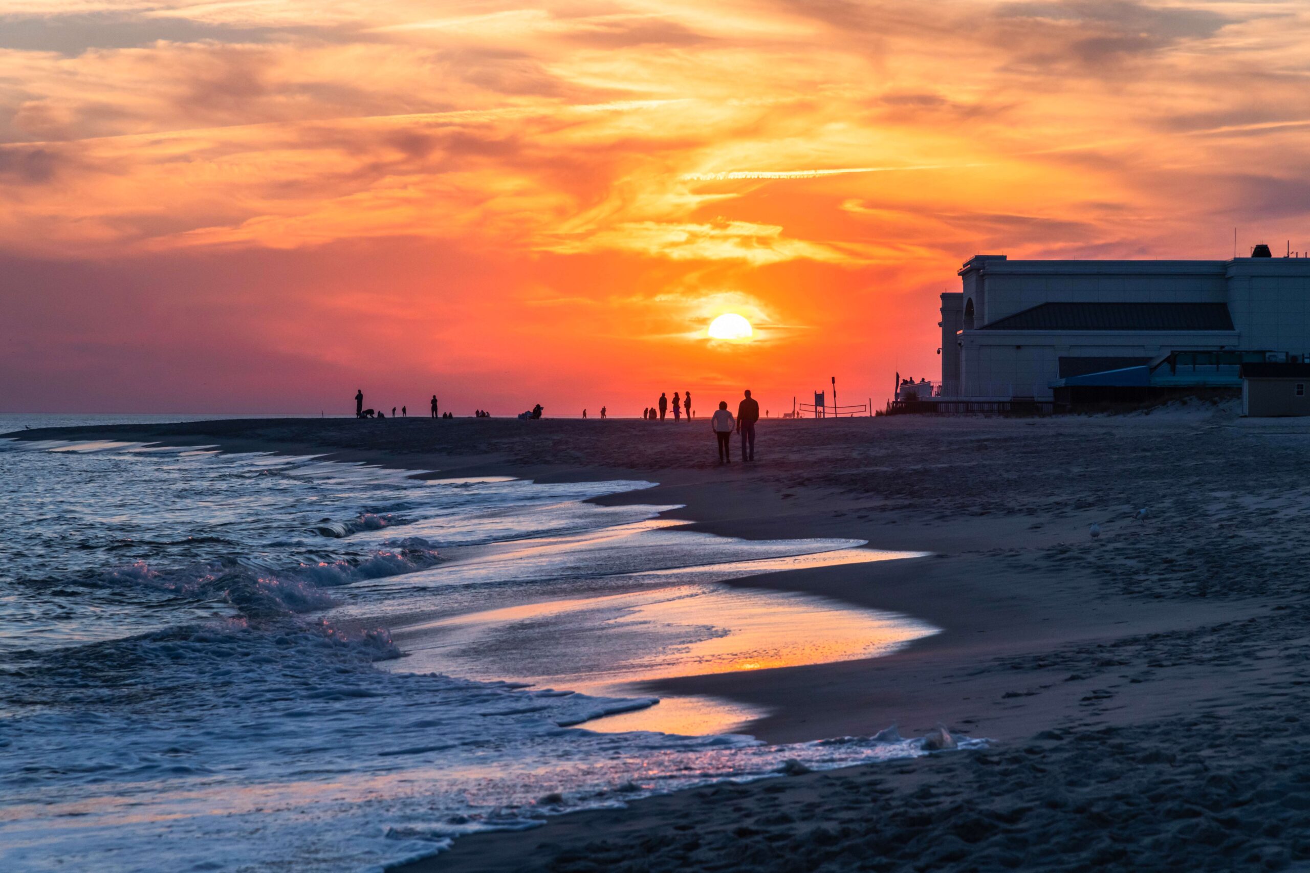 An orange and pink sunset in the sky with groups of people watching at the beach with the ocean rushing into the sand and Convention Hall in the distance