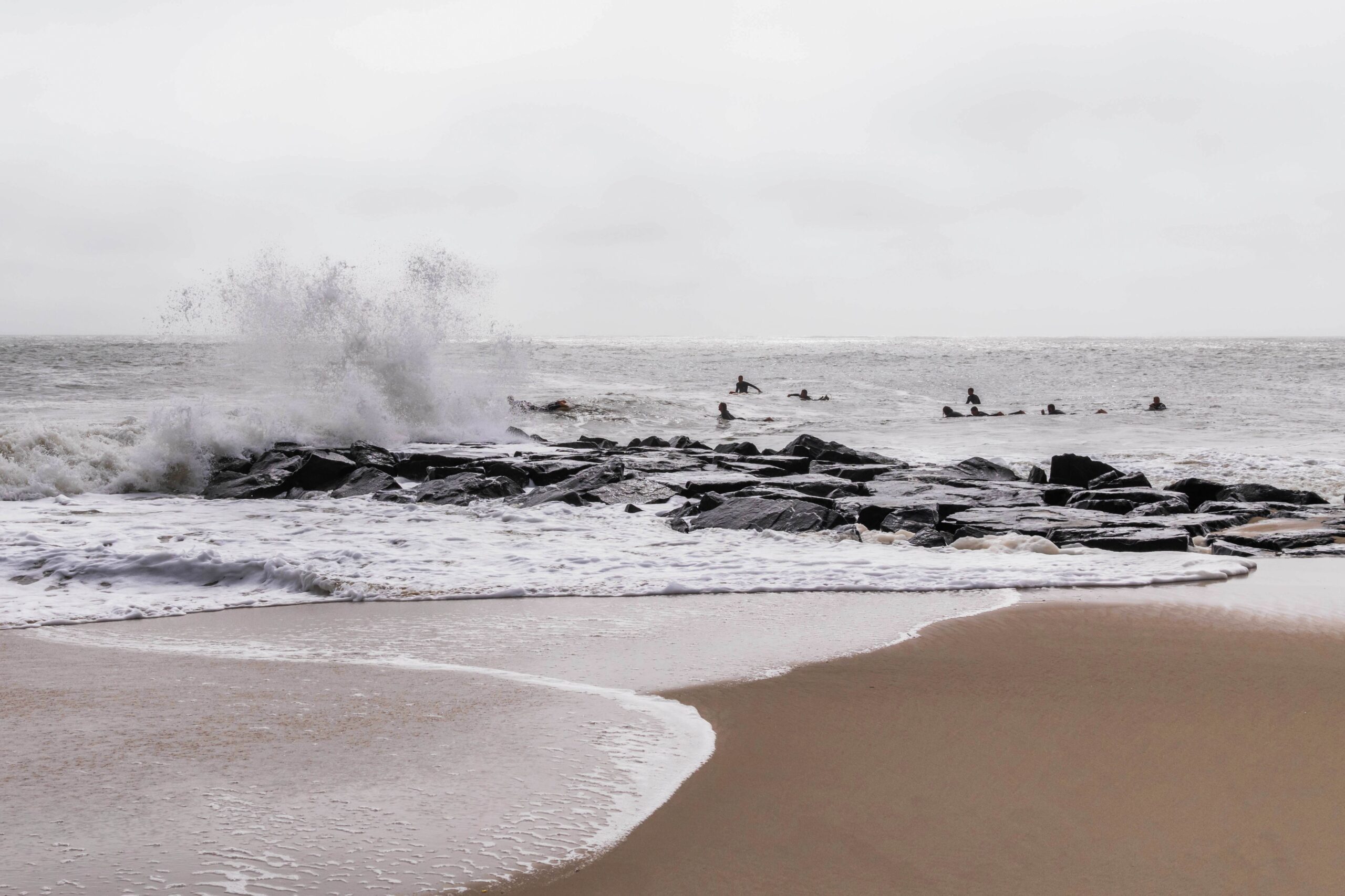 Surfers out in the ocean with a wave crashing on the rocks on a cloudy day
