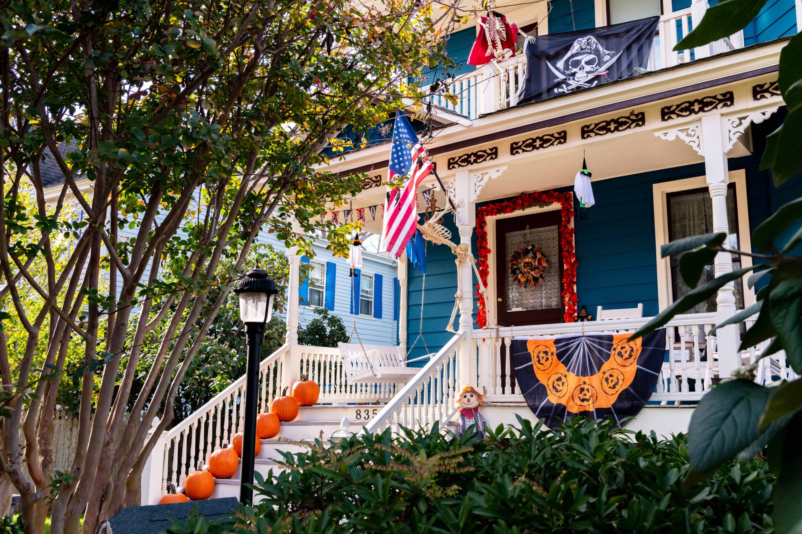 Pumpkins, skeletons, and other halloween decorations on the porch of a teal Victorian home on a sunny day