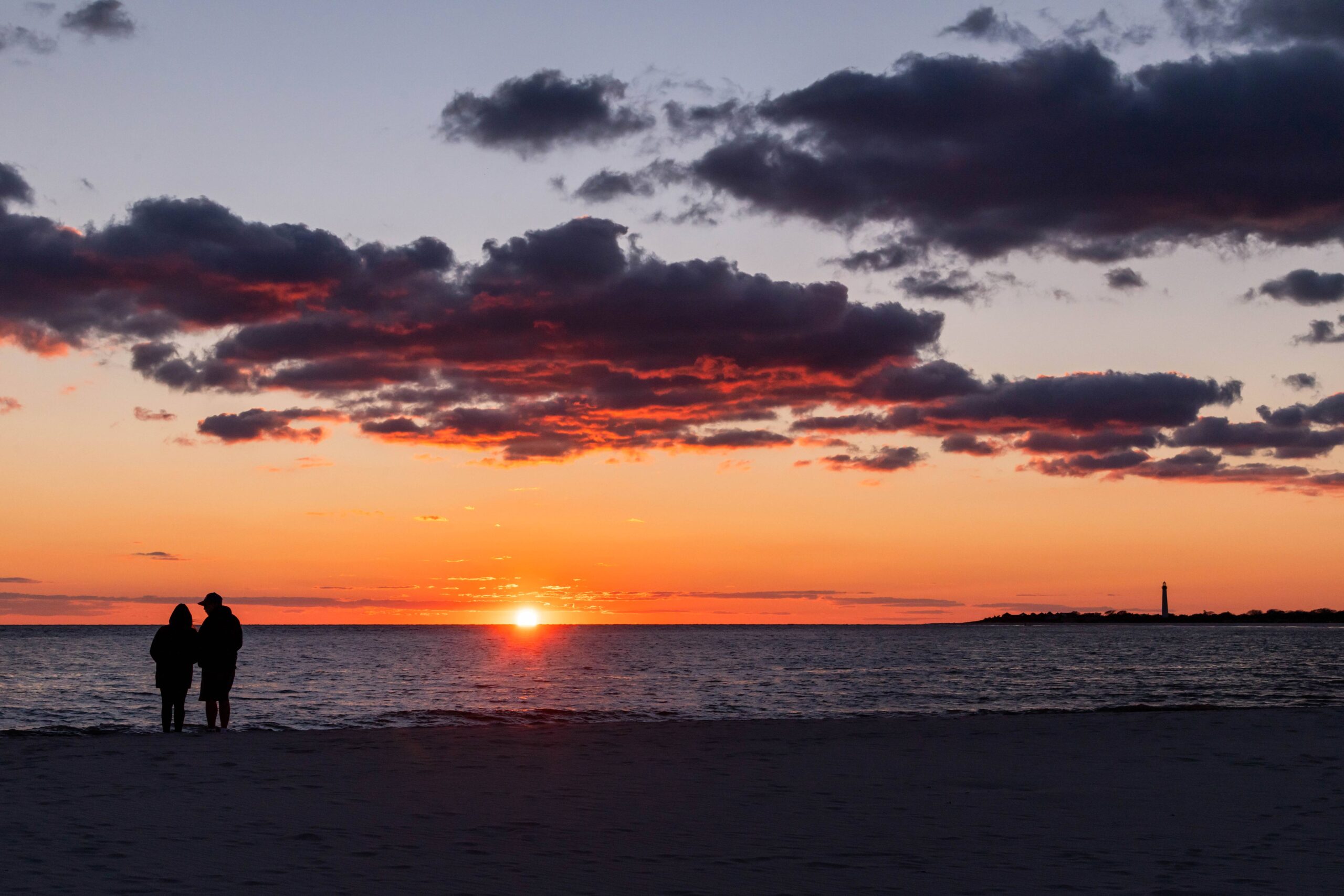 Two people silhouetted watching the sunset with purple and red clouds in the sky and the Cape May lighthouse in the distance