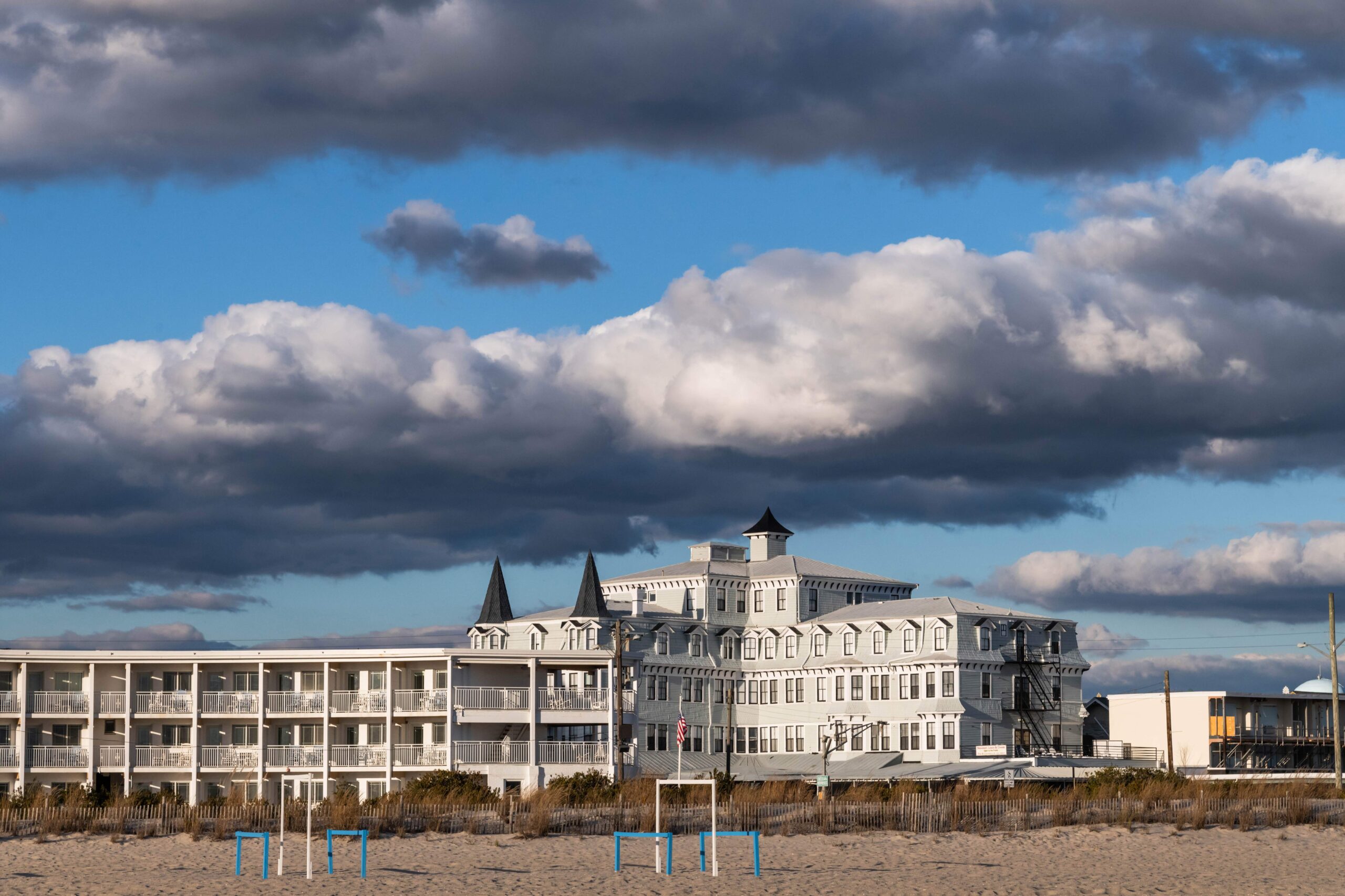 The Inn of Cape May, a white and gray Victorian styled bed and breakfast, with the beach in the foreground and white and gray puffy clouds in a blue sky