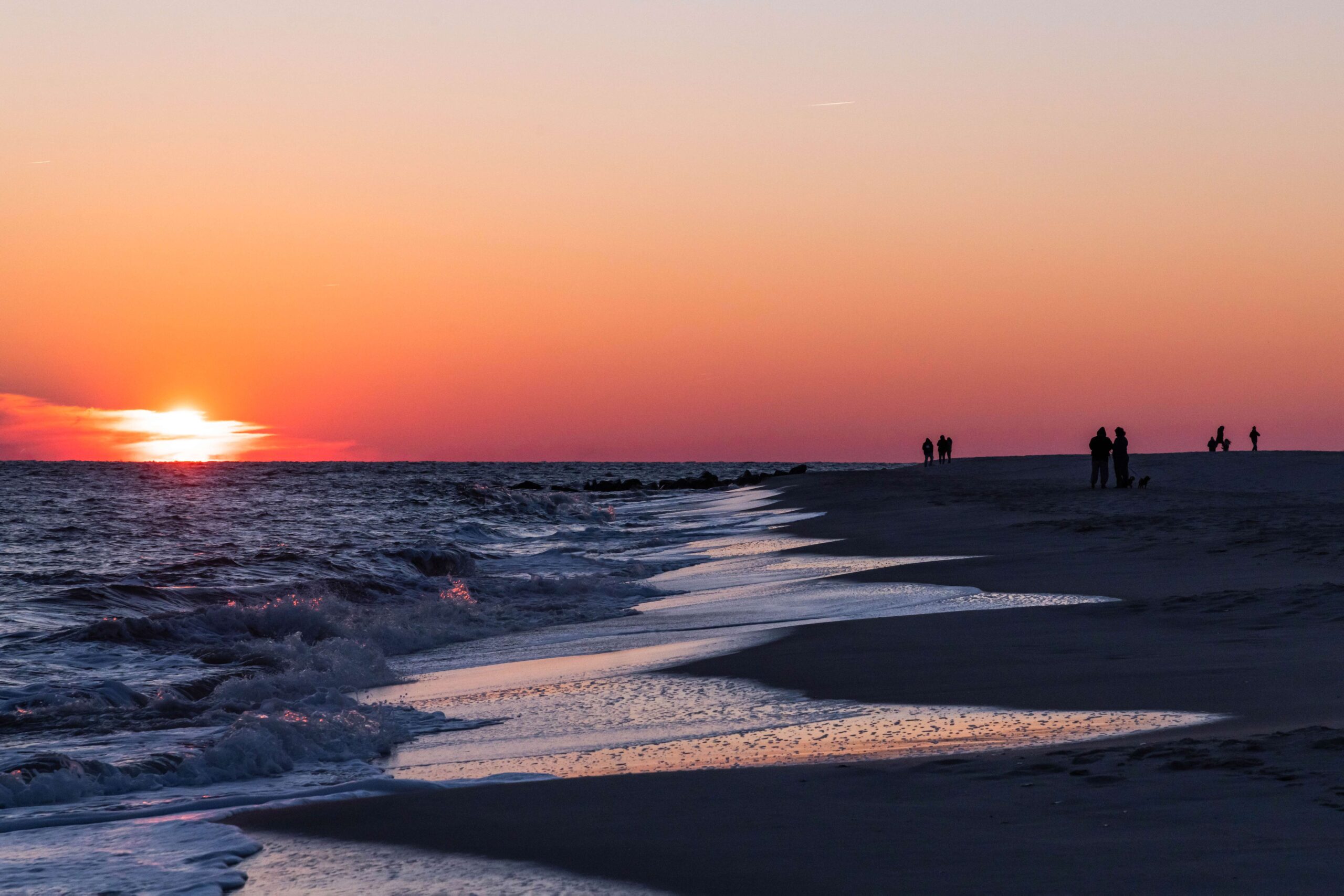 People at the beach by the ocean watching the sun set behind some thin clouds at the horizon with a clear pink and orange sky