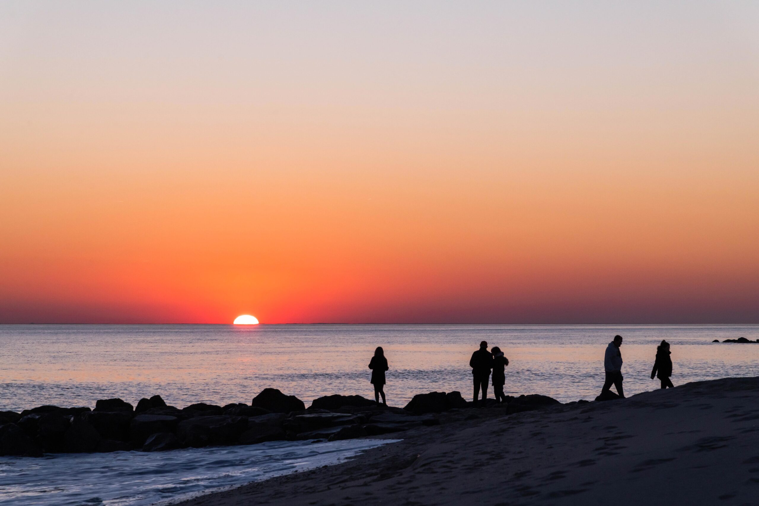 People walking on the beach and on the rocks by the ocean watching the sun dip below the horizon line with pink coloring a clear sky