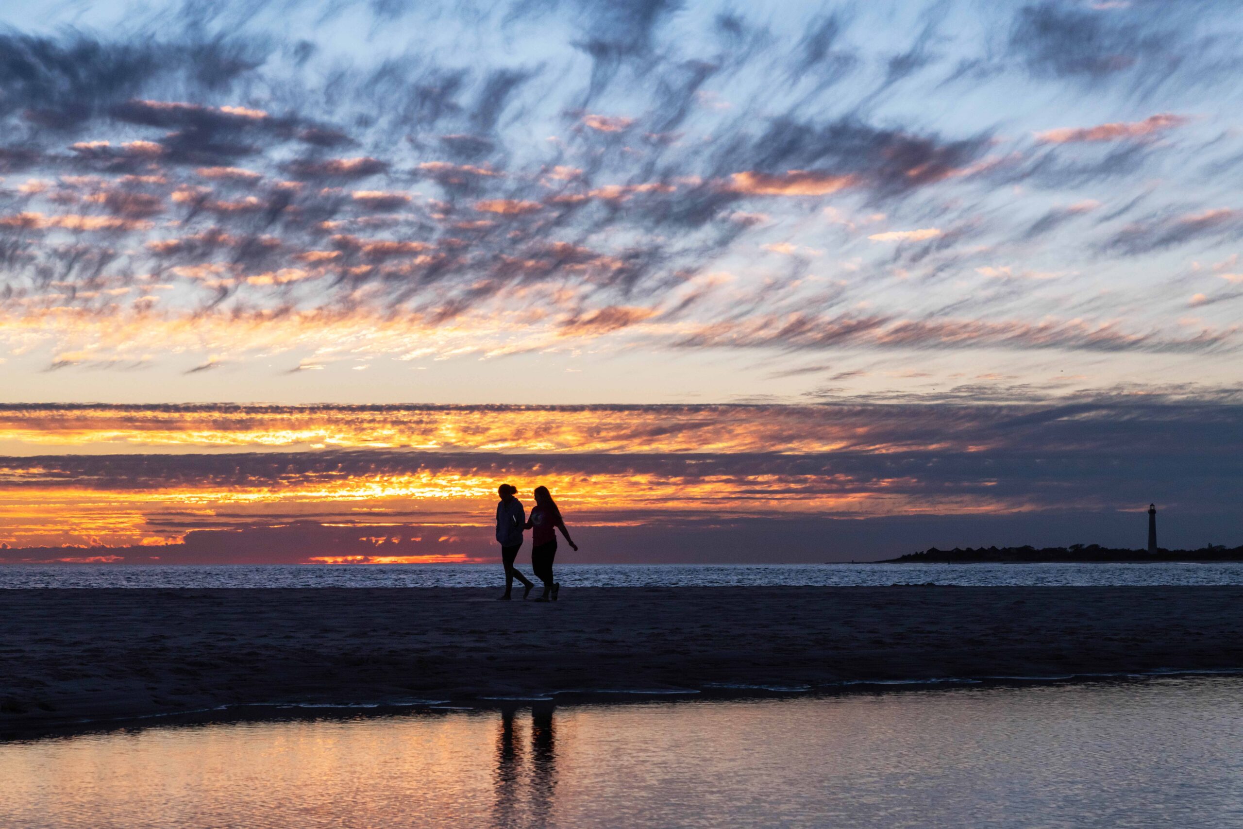 Two people walking on the beach with purple, orange, and pink clouds at the horizon line at sunset. The Cape May Lighthouse is in the distance, and the sunset and people walking are reflected in a pool of water in the sand