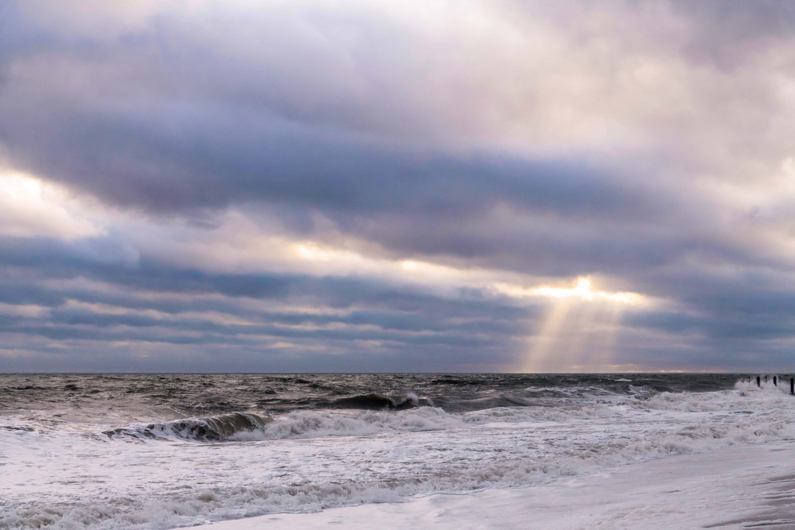 Sunlight streaming through blue and gray muddy dark clouds with the ocean churning and waves crashing on the beach