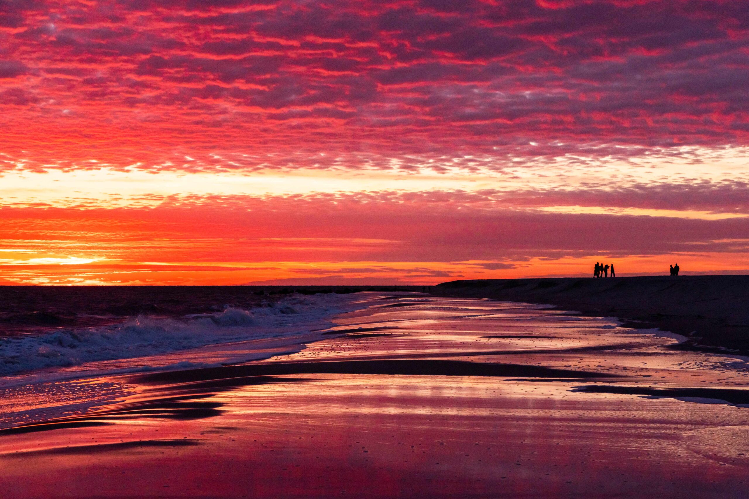 Pink, purple, orange, and yellow bright clouds in the sky at sunset reflected in the ocean and shoreline with waves crashing and people walking in the distance on the beach