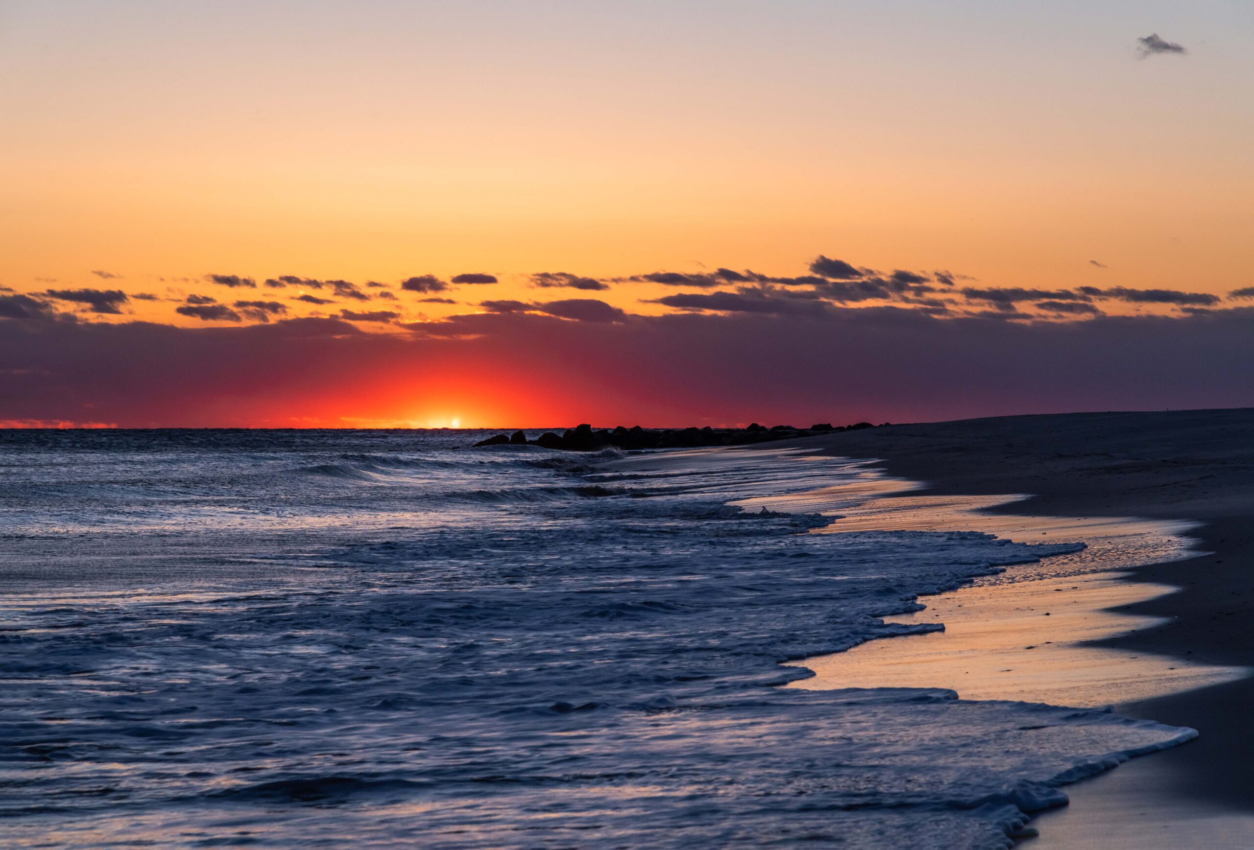 A bright pink spotlight at the horizon where the sun set with purple clouds at the horizon line and the bleu ocean rushing into the beach