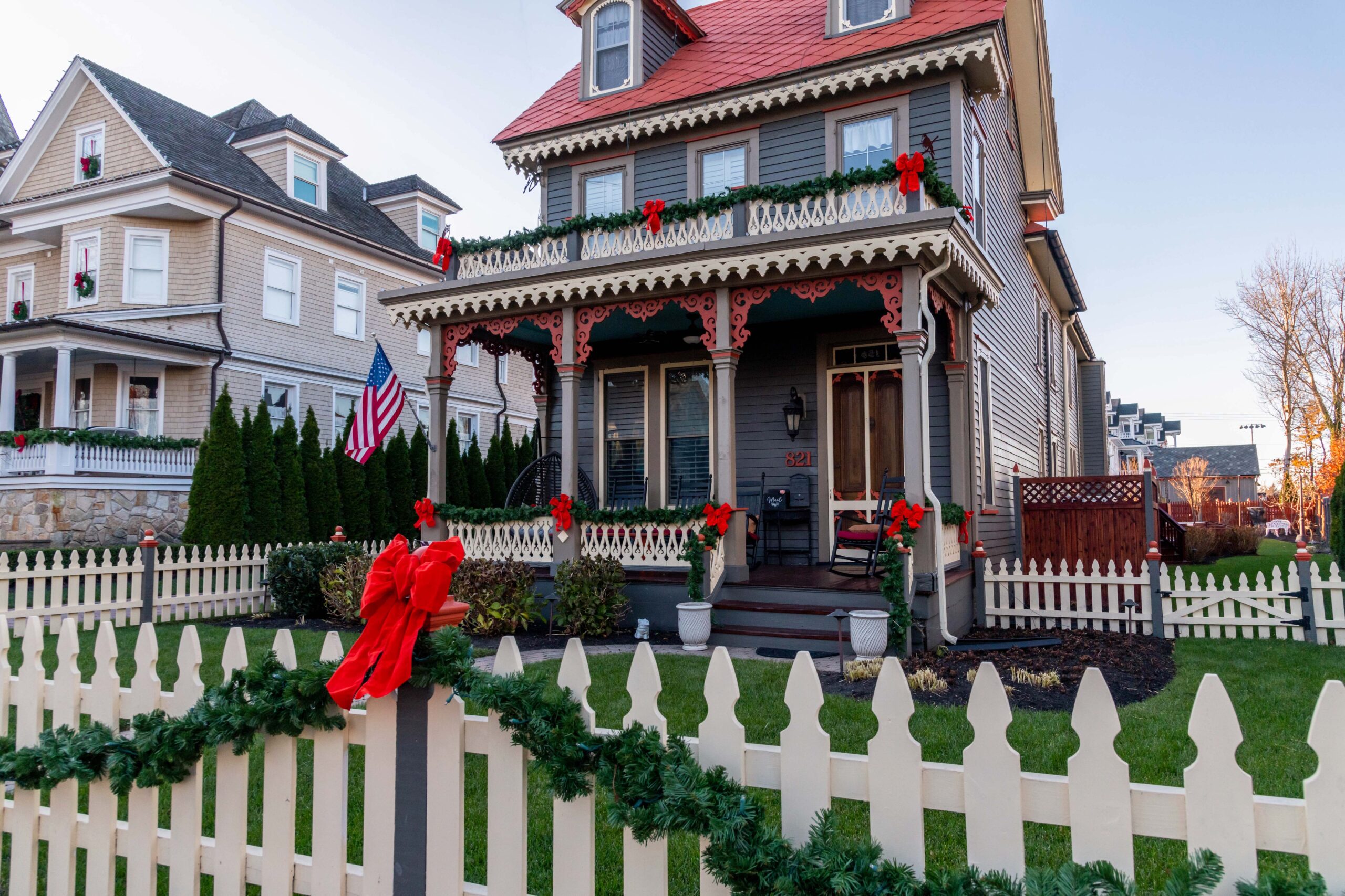 Red bows and green garland hung up on the fence and the porch railings of a red, gray, and pale yellow Victorian style house with a clear blue sky