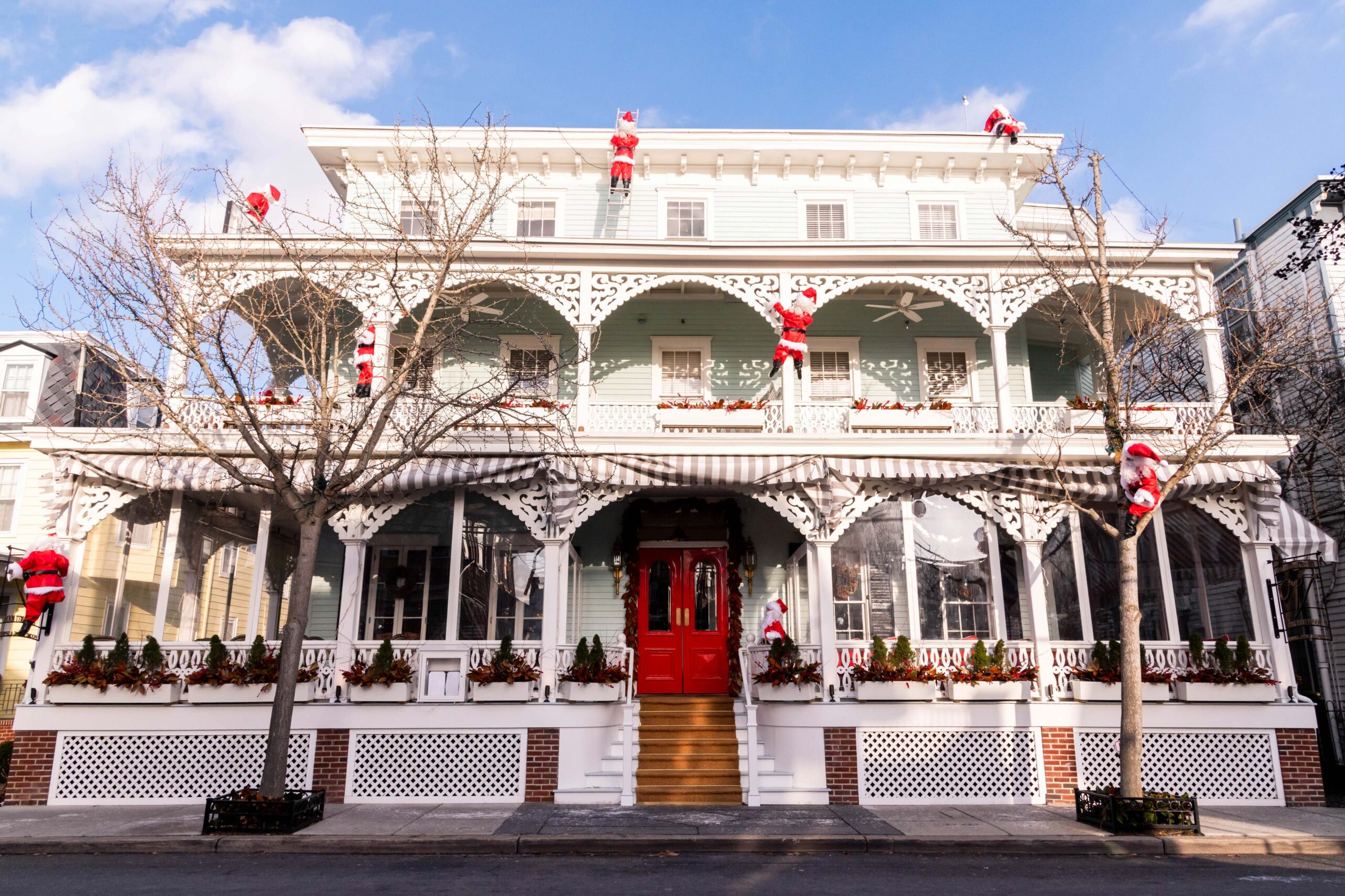 Santa's climbing on porch railings and on the roof of Virginia Hotel, a pastel blue and white Victorian bed and breakfast. The sky is blue with puffy white clouds and the sun is shining on the Virginia Hotel.