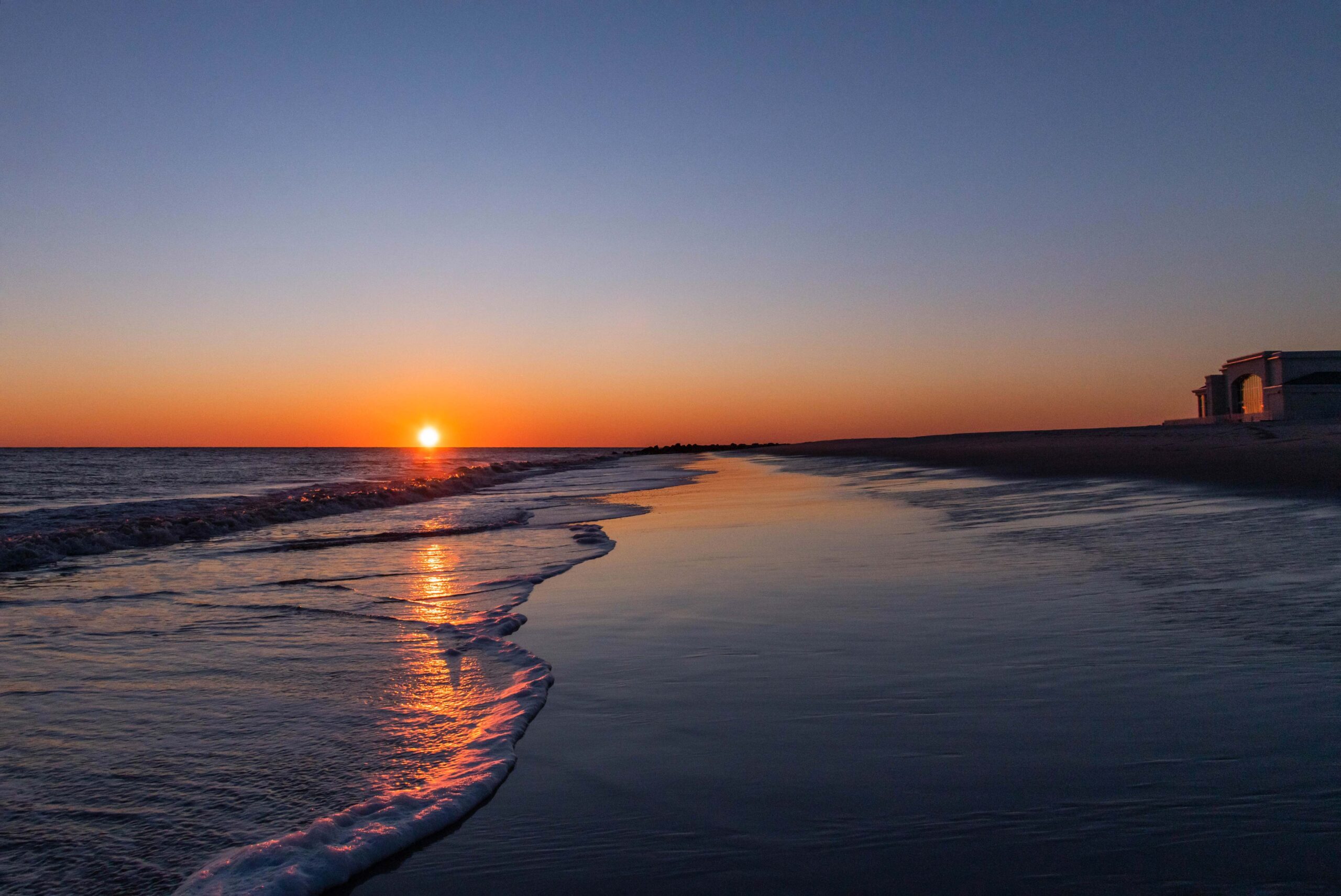 Sun setting with the ocean coming into the shore and a bright clear sky. Convention Hall is in the distance on the horizon line, and sunlight is shining on the windows