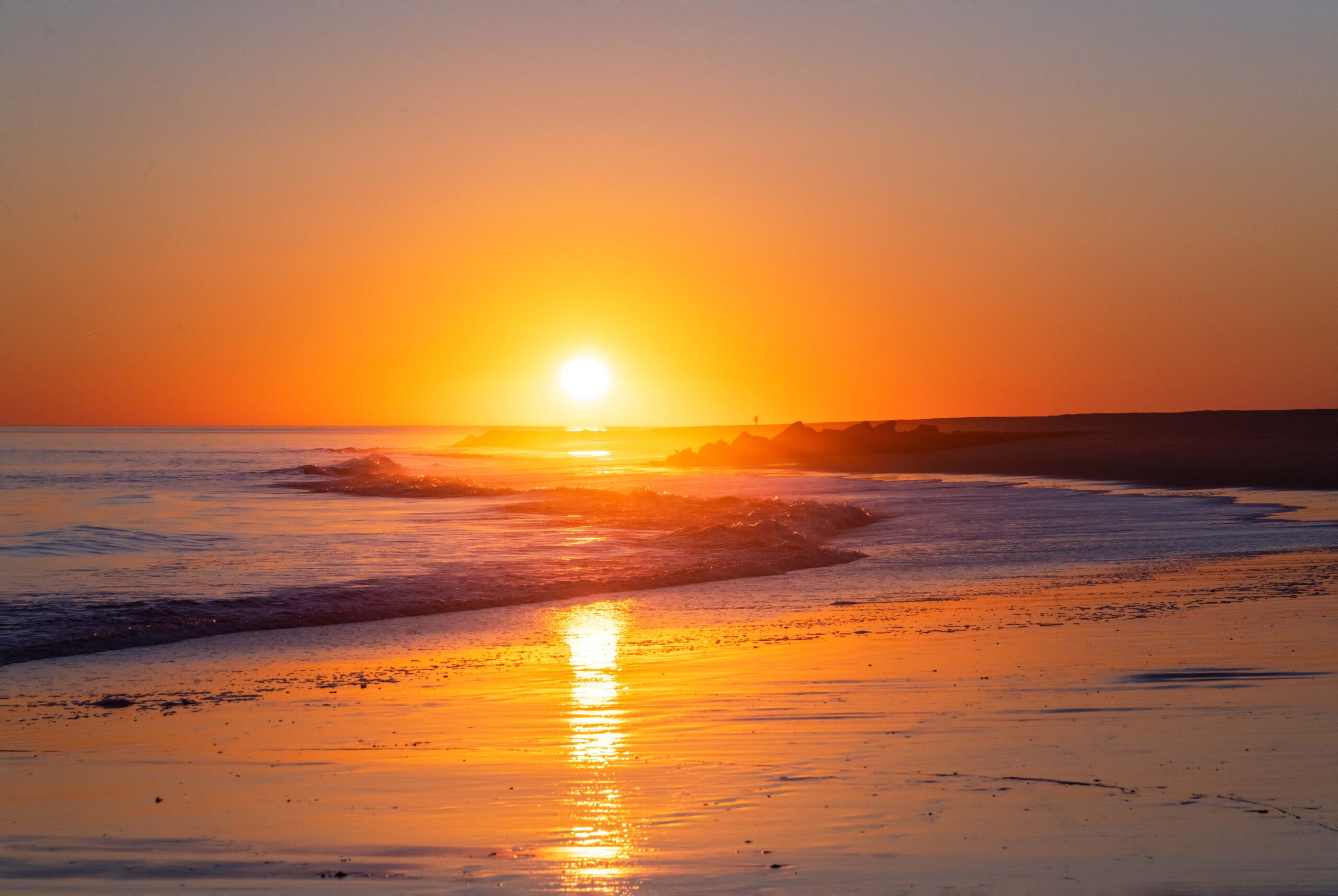 A bright yellow sun setting with a clear sky. The ocean is rolling into the shore and sunlight is reflected on the shoreline