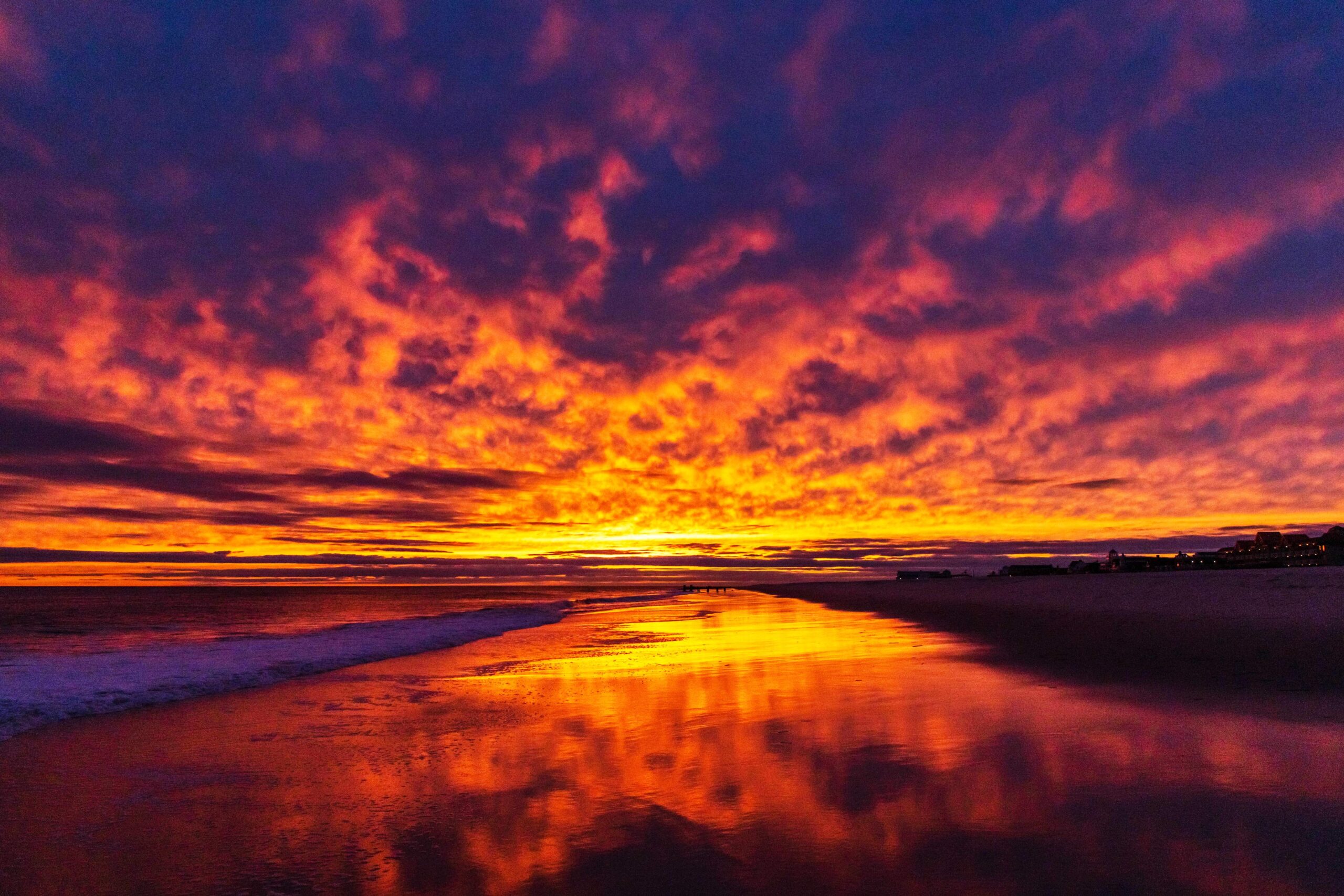 Brilliant purple, pink, orange, and yellow clouds in the sky at sunset, reflected in the ocean and the shoreline at the beach