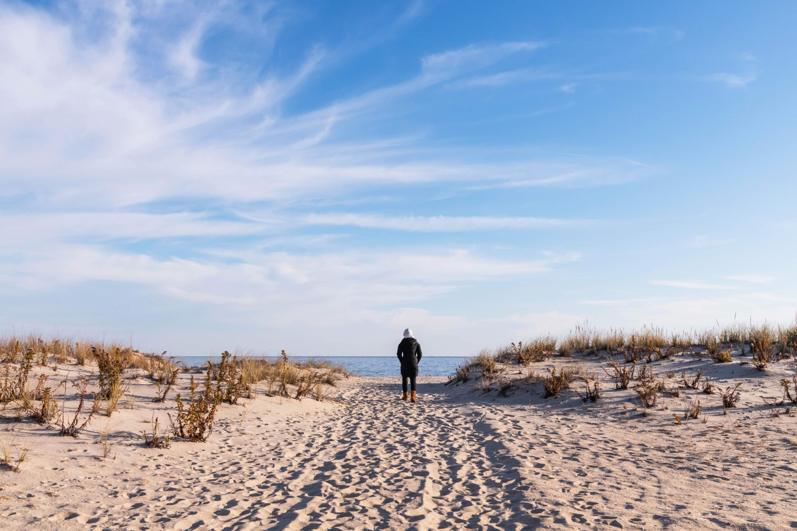 A person bundled up walking down a path on the beach with dunes on either side. There are wispy white clouds in the blue sky and the ocean is in the distance