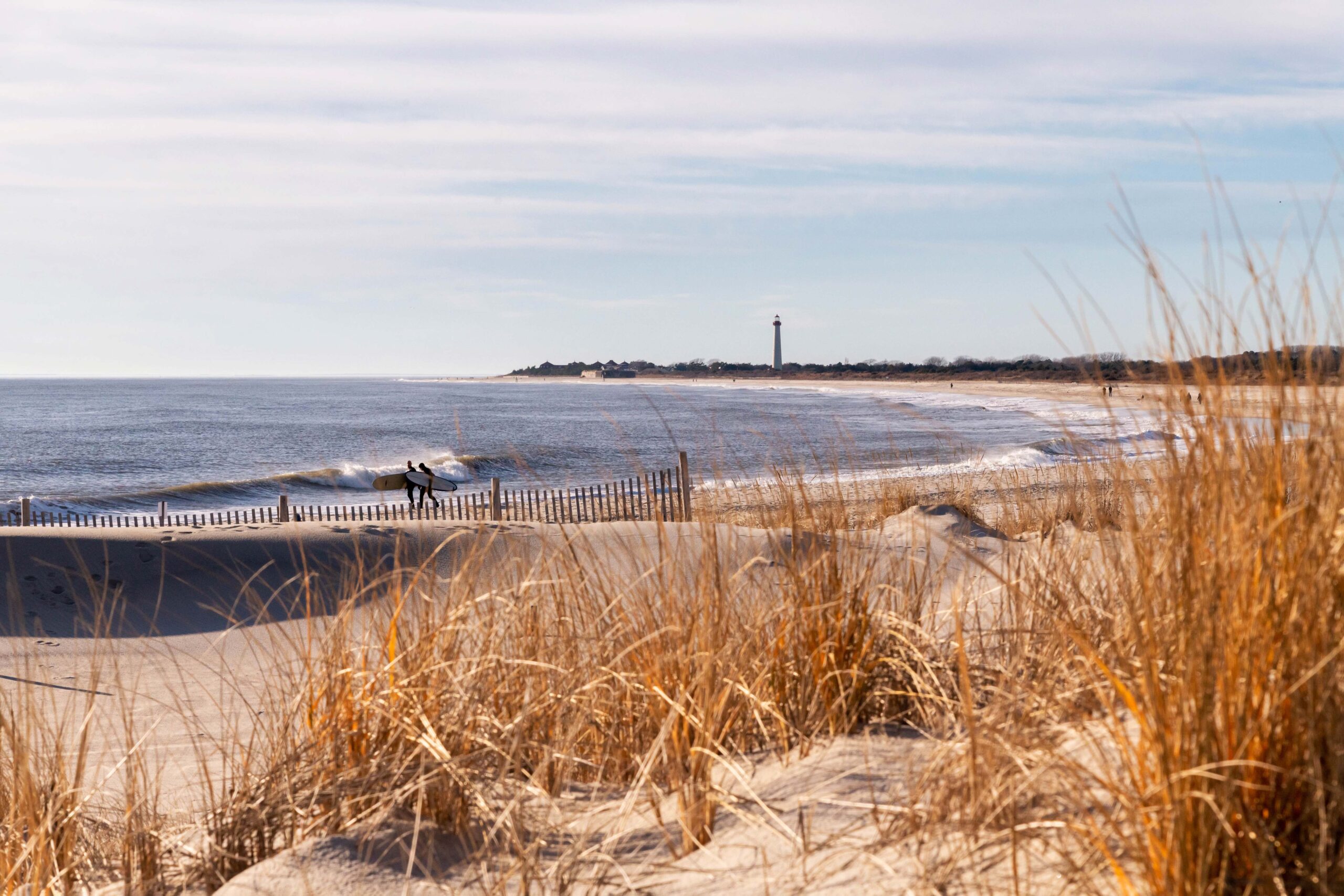 Beach dunes blurred out in the foreground with two surfers walking by the ocean. Waves are crashing and the Cape May Lighthouse is in the distance 