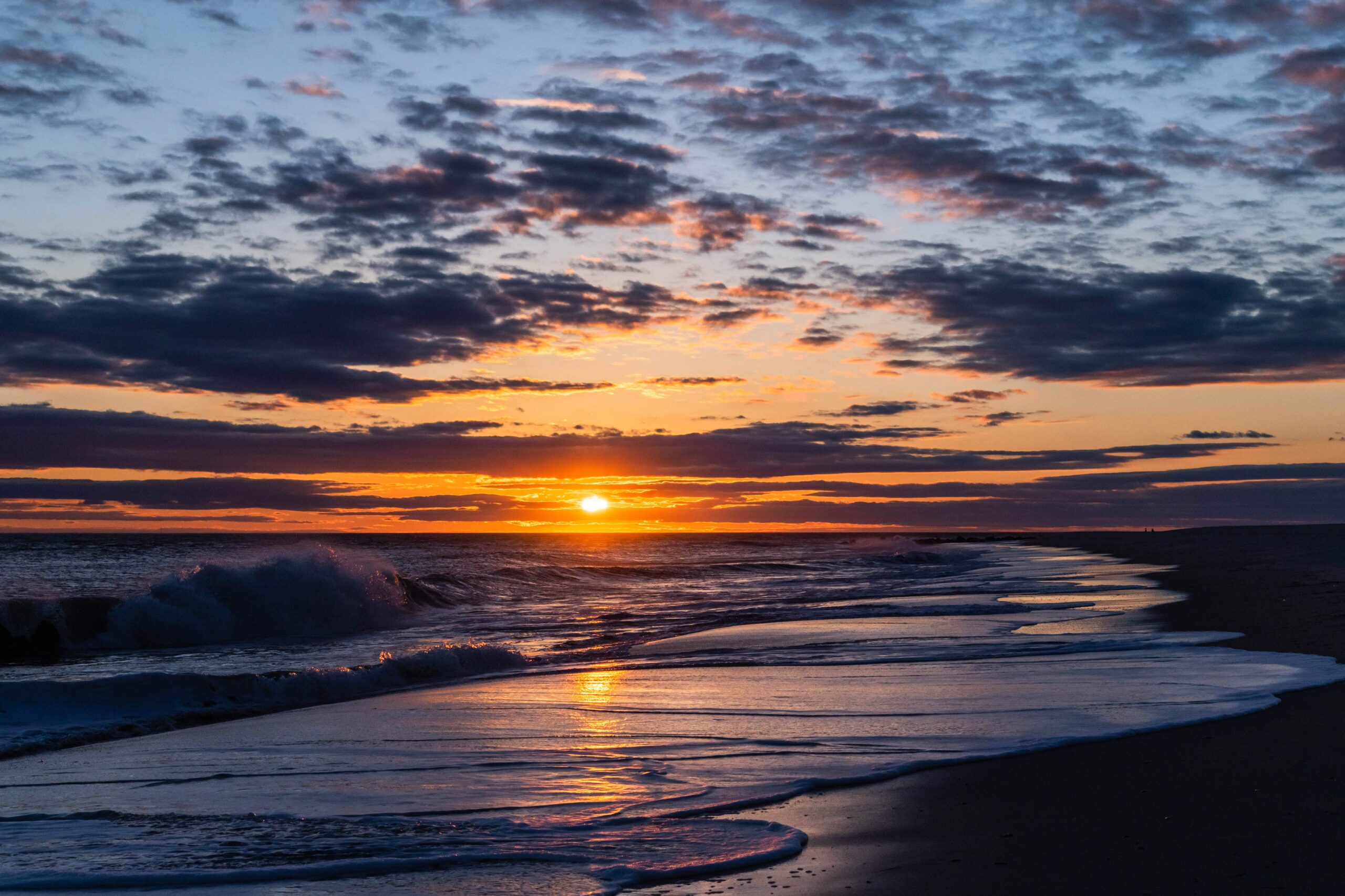 Thin purple clouds in the sky with the sun setting and waves crashing at the beach