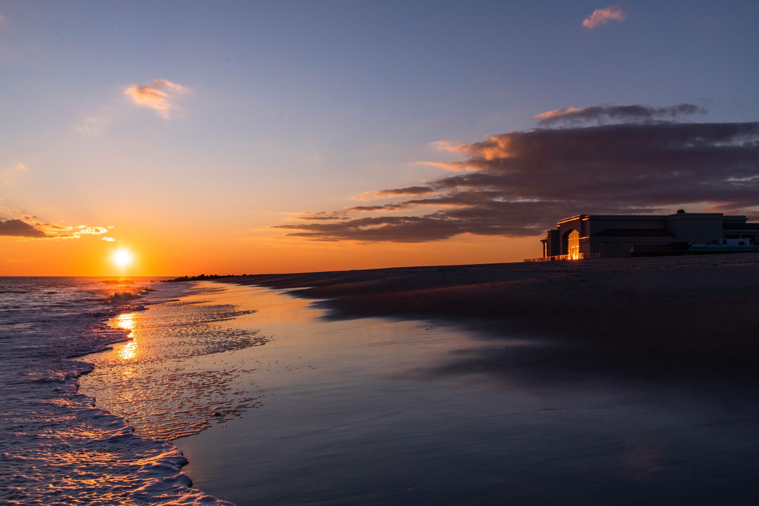 Sun setting with a few clouds in the sky, the ocean rushing into the beach, and Convention Hall in the distance