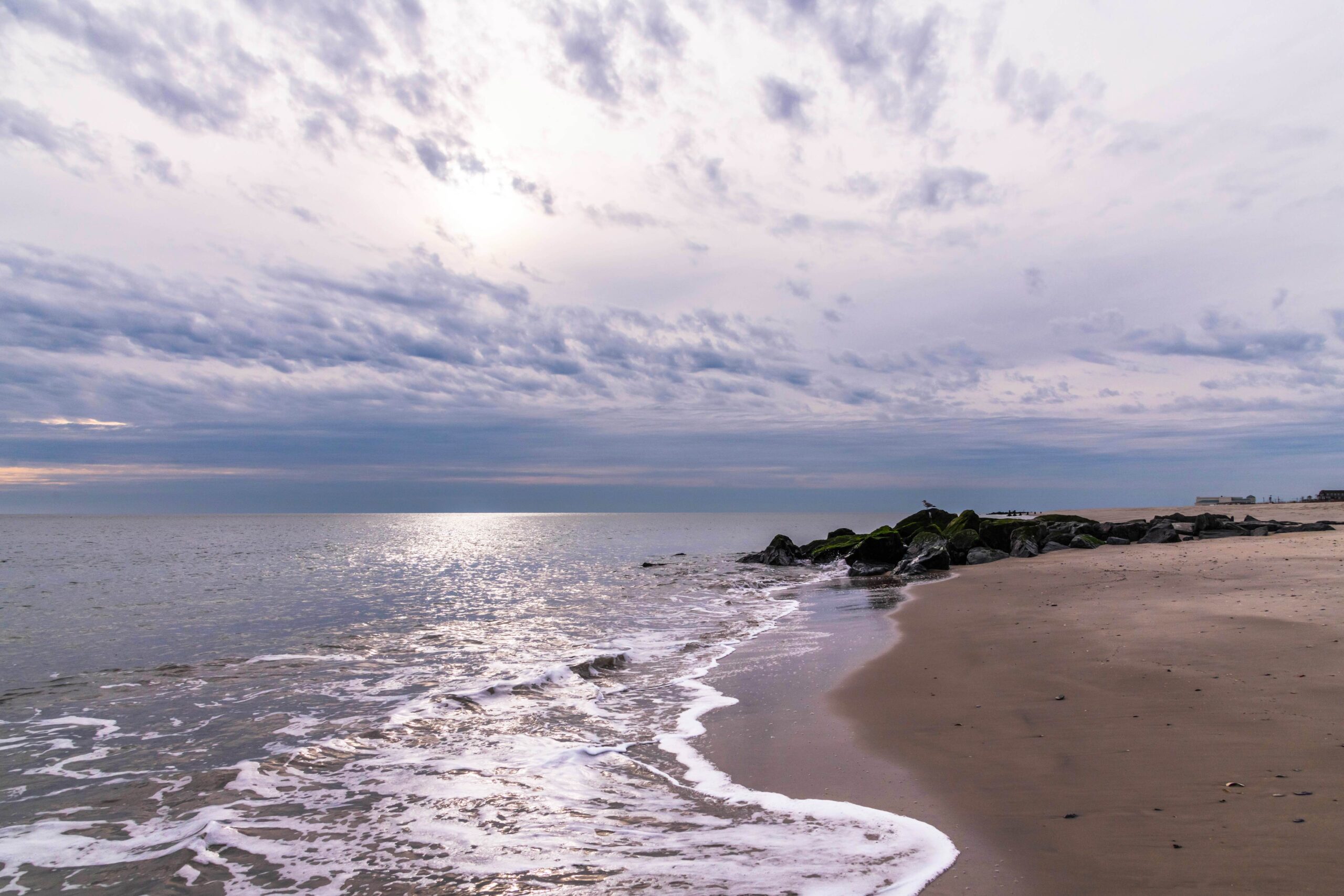 Blue and gray clouds in the sky with the ocean coming into the shoreline and a seagull perched on a rock jetty