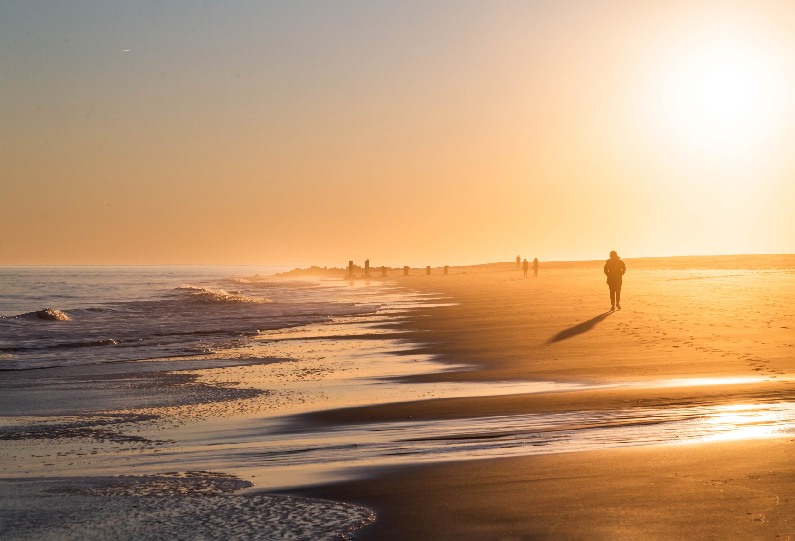 A person walking on the beach by the ocean with a clear sky and the sun shining intensely causing a golden glow on the beach