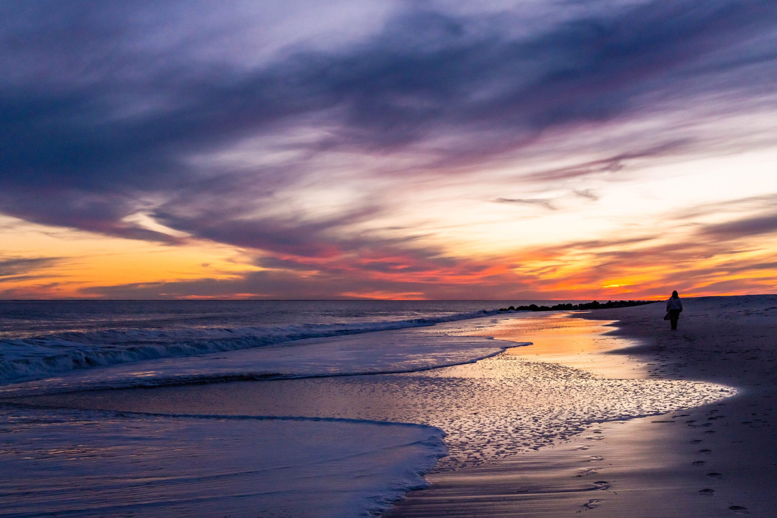 Purple, Pink, and orange wispy clouds in the sky at sunset with waves coming into the shoreline and someone walking by the ocean