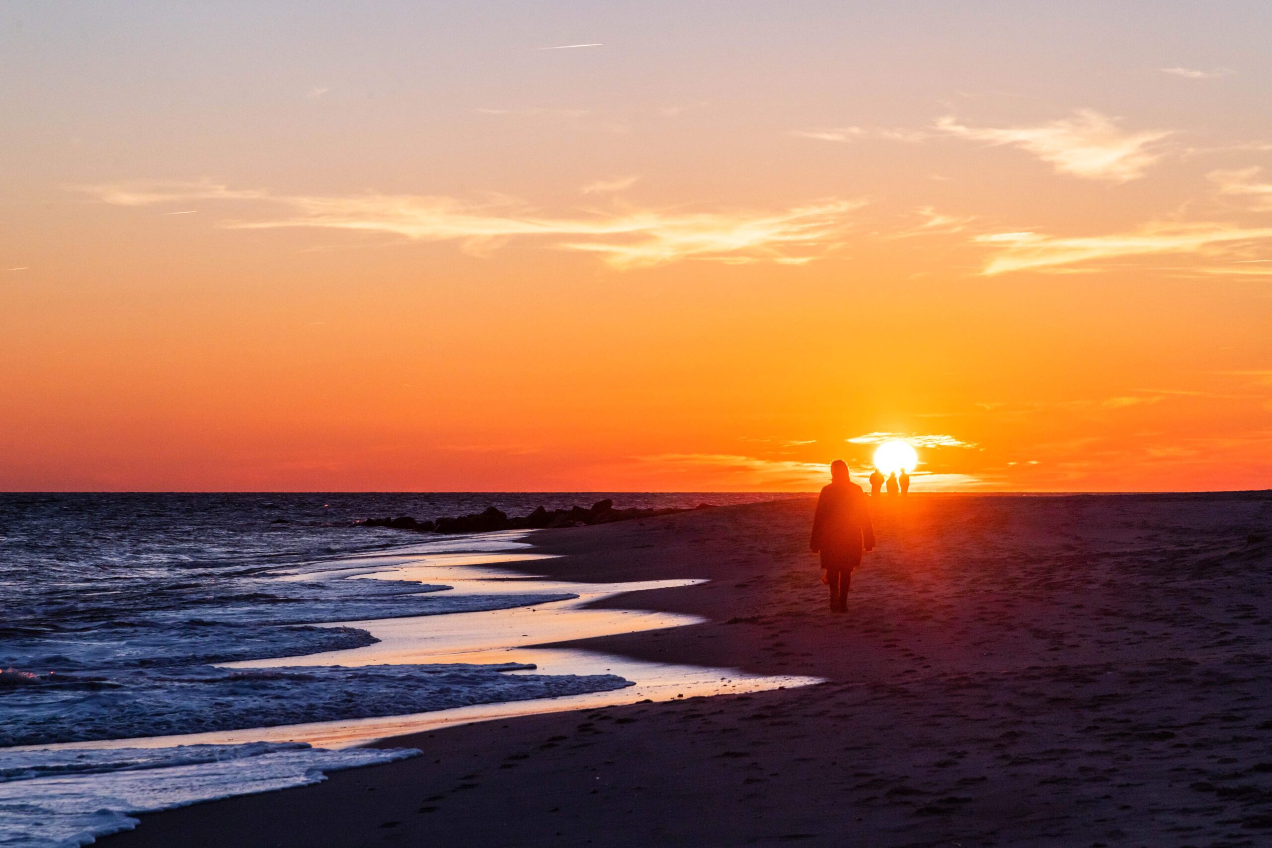 The sun setting behind three people in the distance while someone walks along the ocean