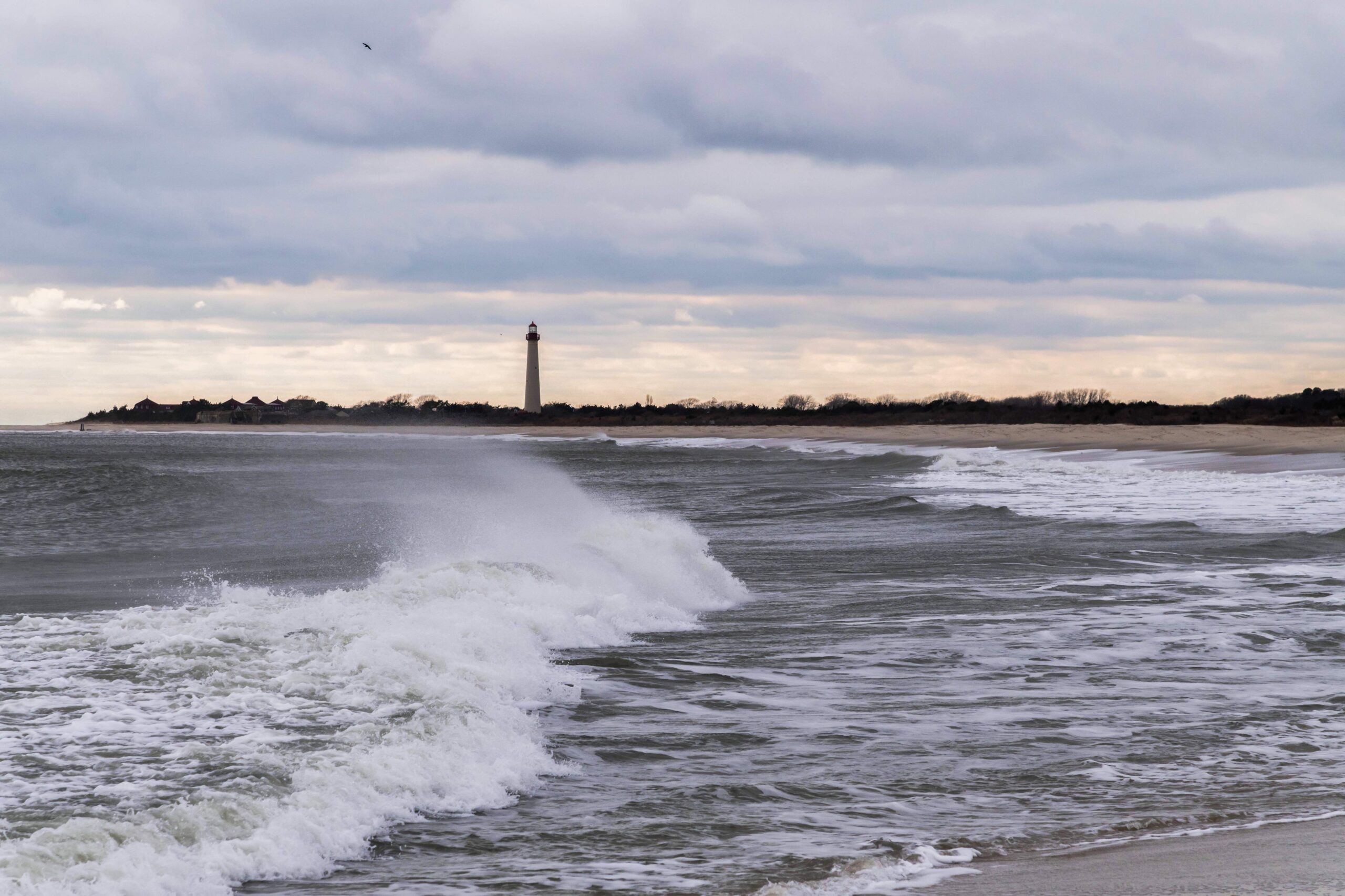 Waves crashing and spraying with the Cape May Lighthouse in the distance and clouds in the sky