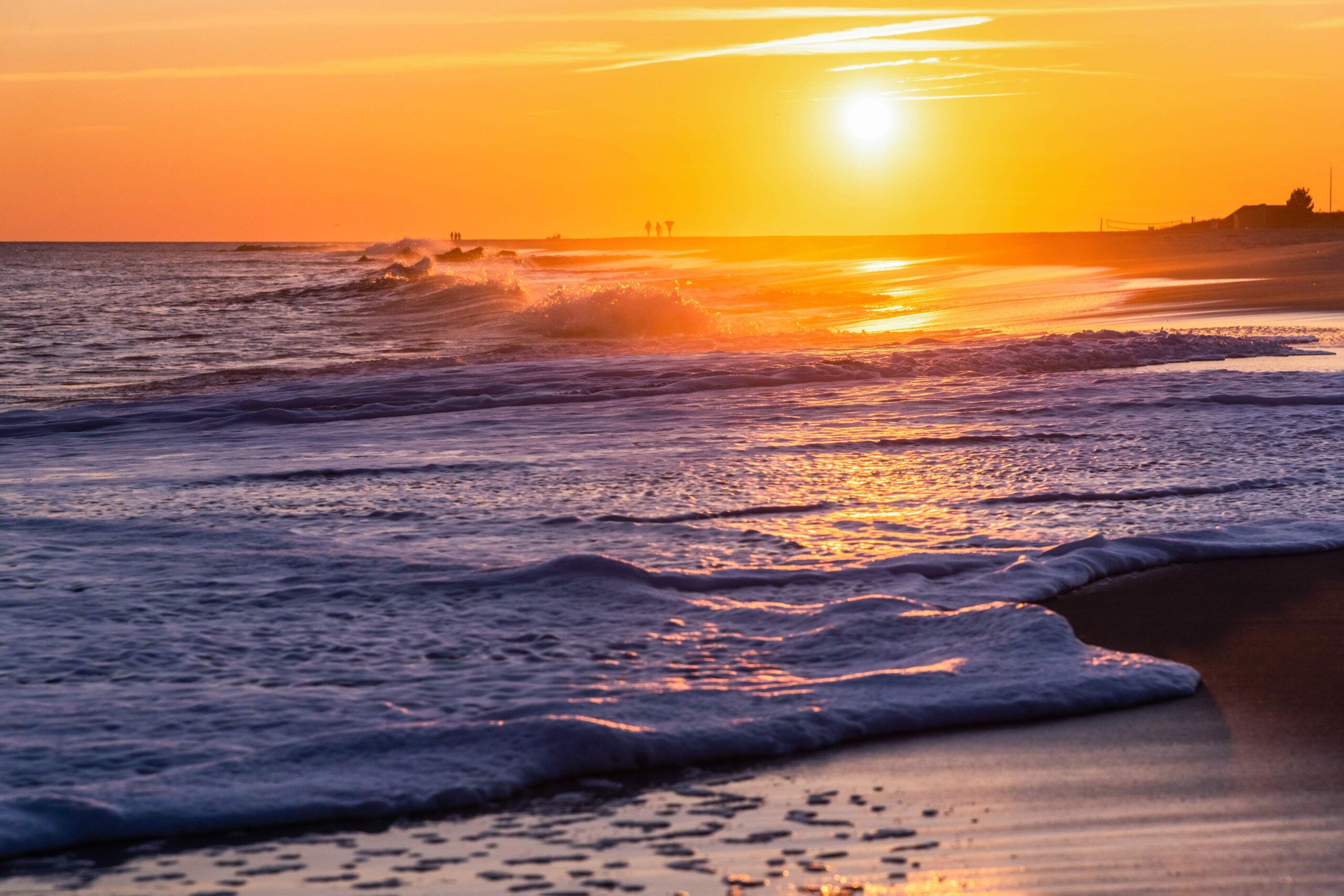 A close up of the ocean rushing into the beach with the sun setting in a clear orange sky and silhouettes of people walking on the beach in the distance