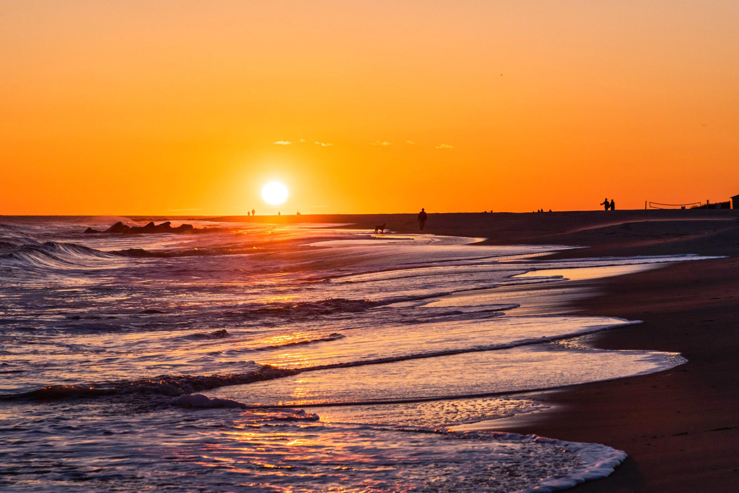 Waves rushing into the shoreline while people walk on the beach in the distance at sunset with a clear orange sky