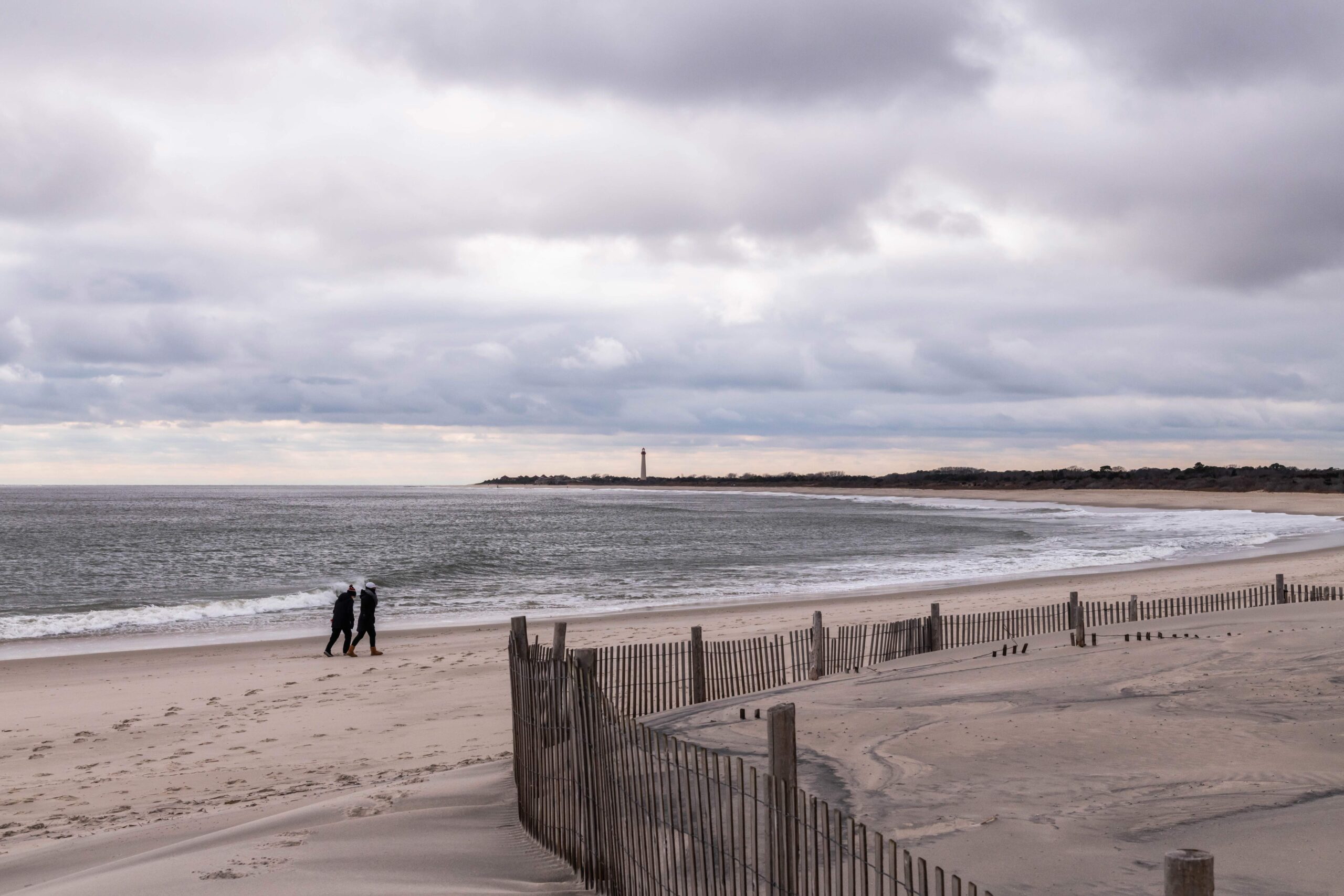 Two people walking on the beach towards the lighthouse with gray puffy clouds in the sky and a beach fence in the foreground