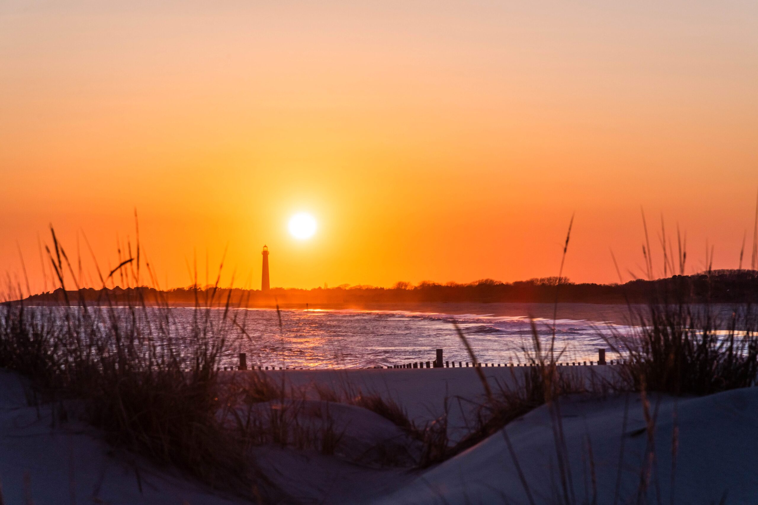 The sun setting by the lighthouse in the distance through beach dunes