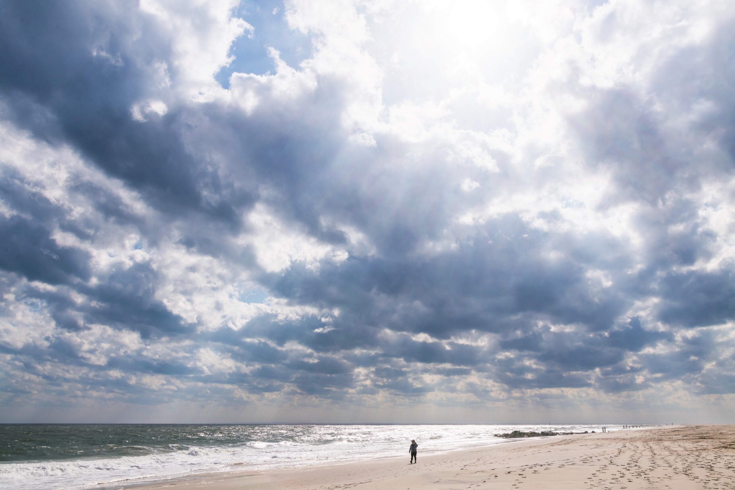 Puffy blue and gray clouds in the sky with sun breaking through and someone walking in the distance on the beach