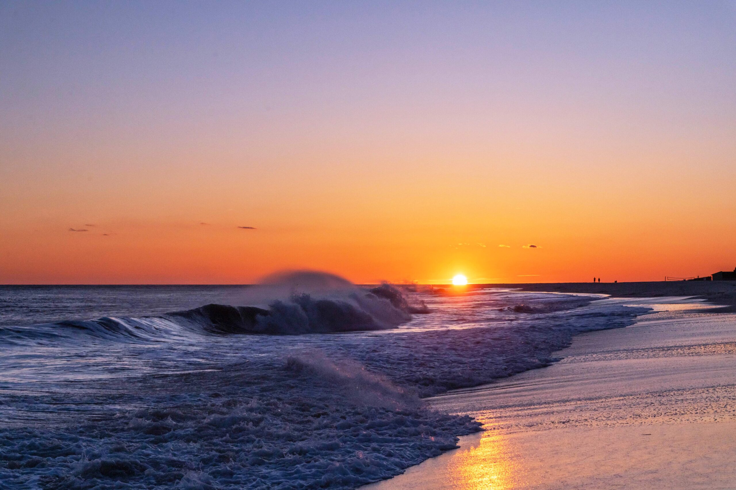 A wave crashing with ocean spray as the sun sets in a clear blue and orange sky