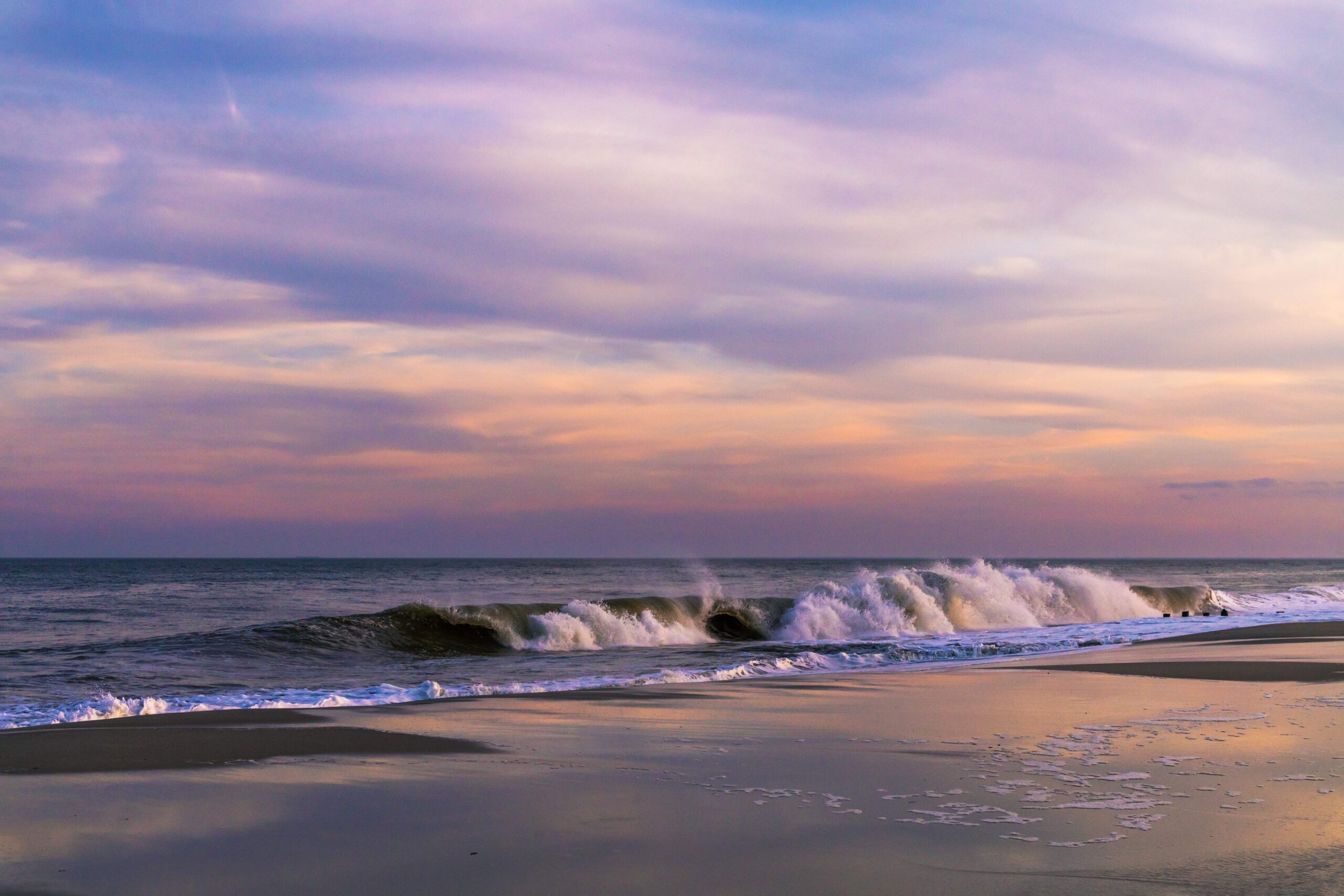 Pink, purple, blue, orange, and yellow clouds in the sky and reflected in the sand with a wave crashing in the ocean