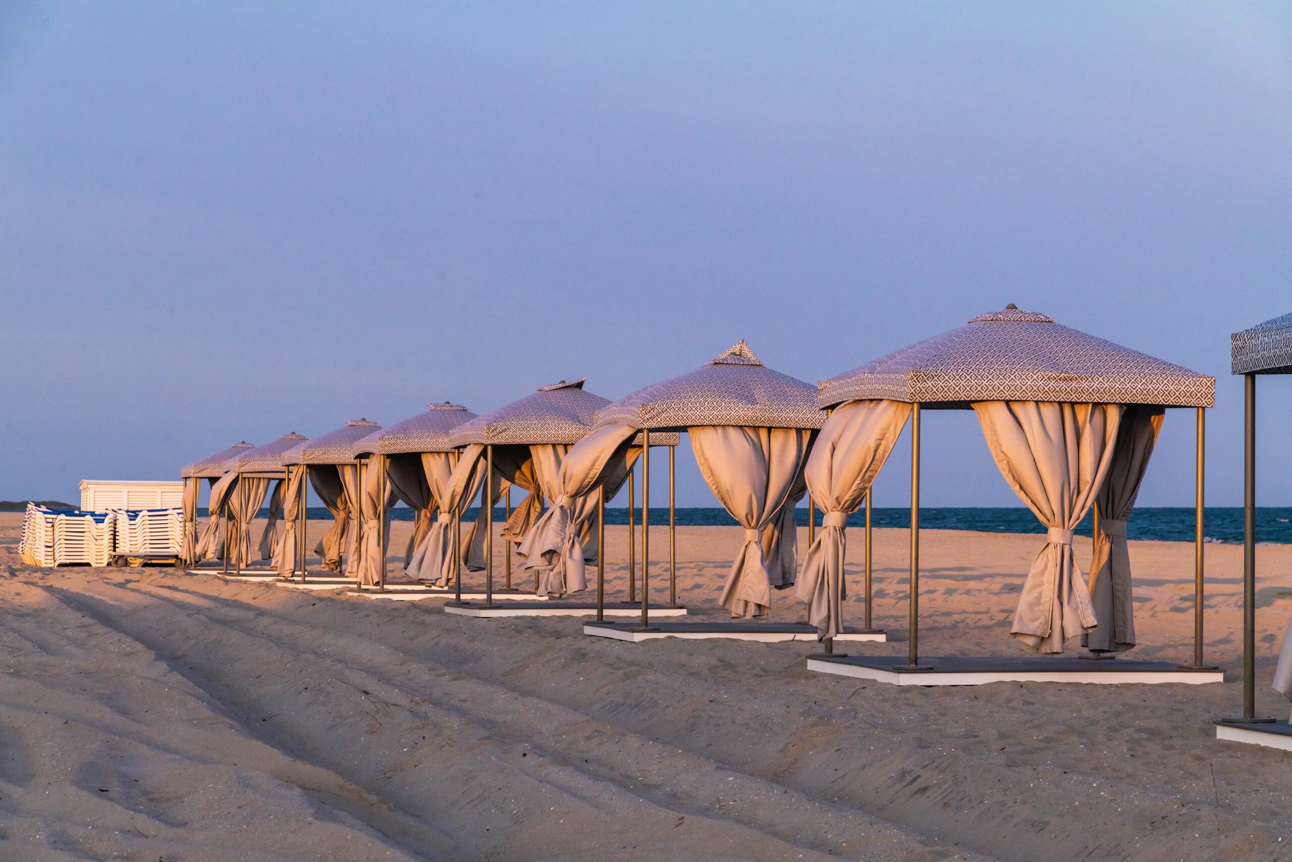 Tan beach cabanas lined up in a row with beach lounge chairs with a blue sky and the ocean in the background at sunset