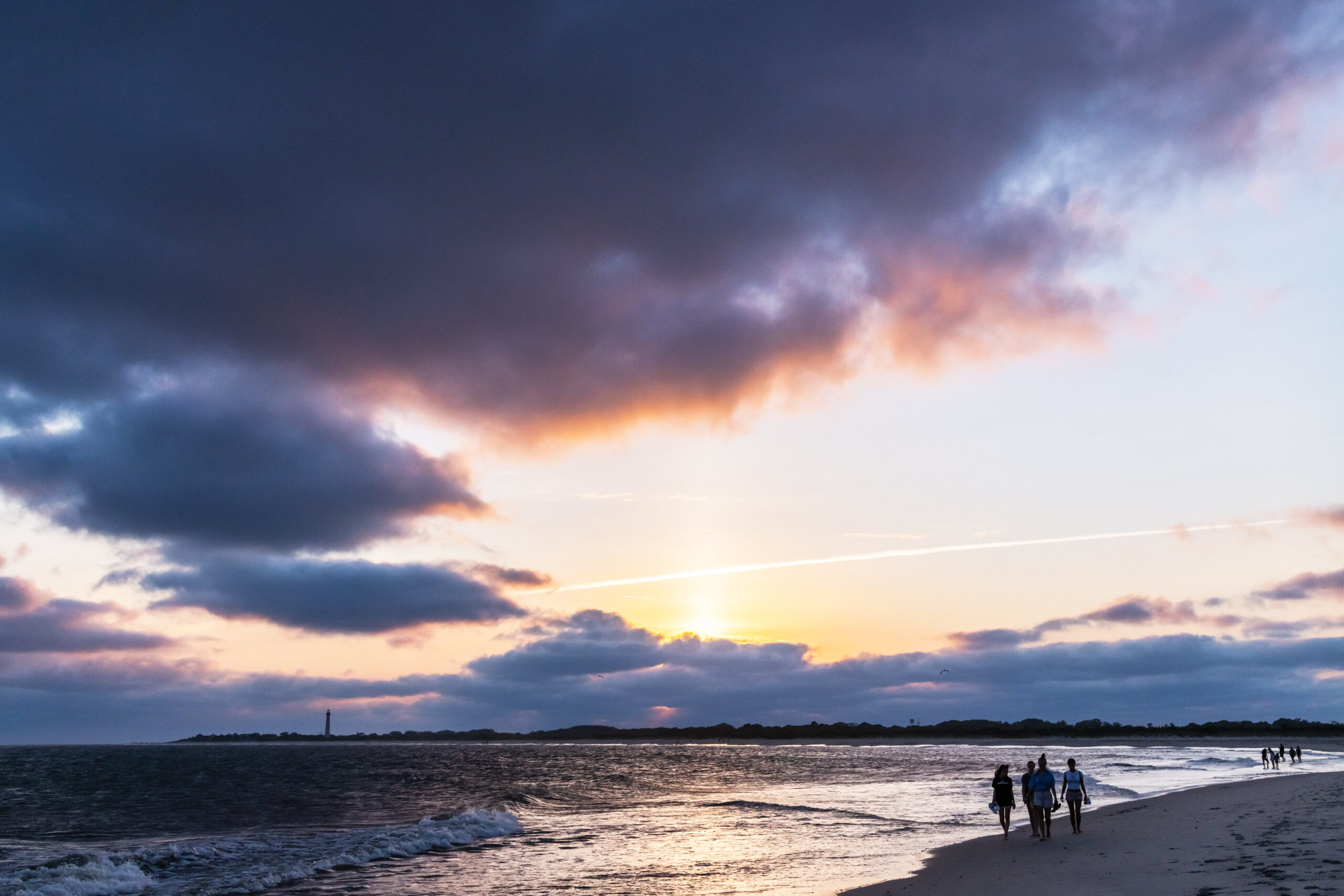 People walking on the beach along the ocean with big purple, blue, and pink clouds in the sky at sunset with the Cape May Lighthouse in the distance