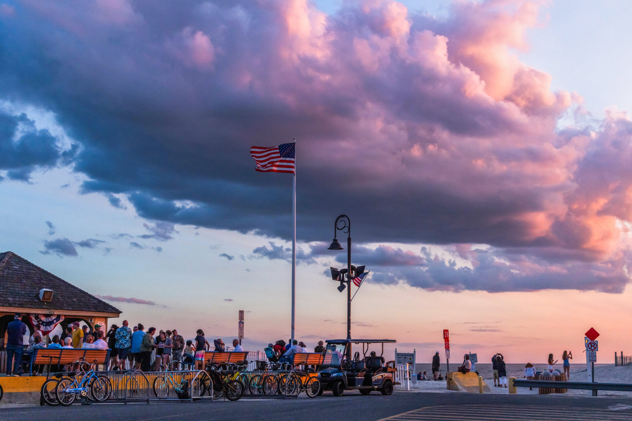 A pink, purple, and blue cloud in the sky at sunset with people gathered at the end of the promenade and an American Flag blowing in the wind