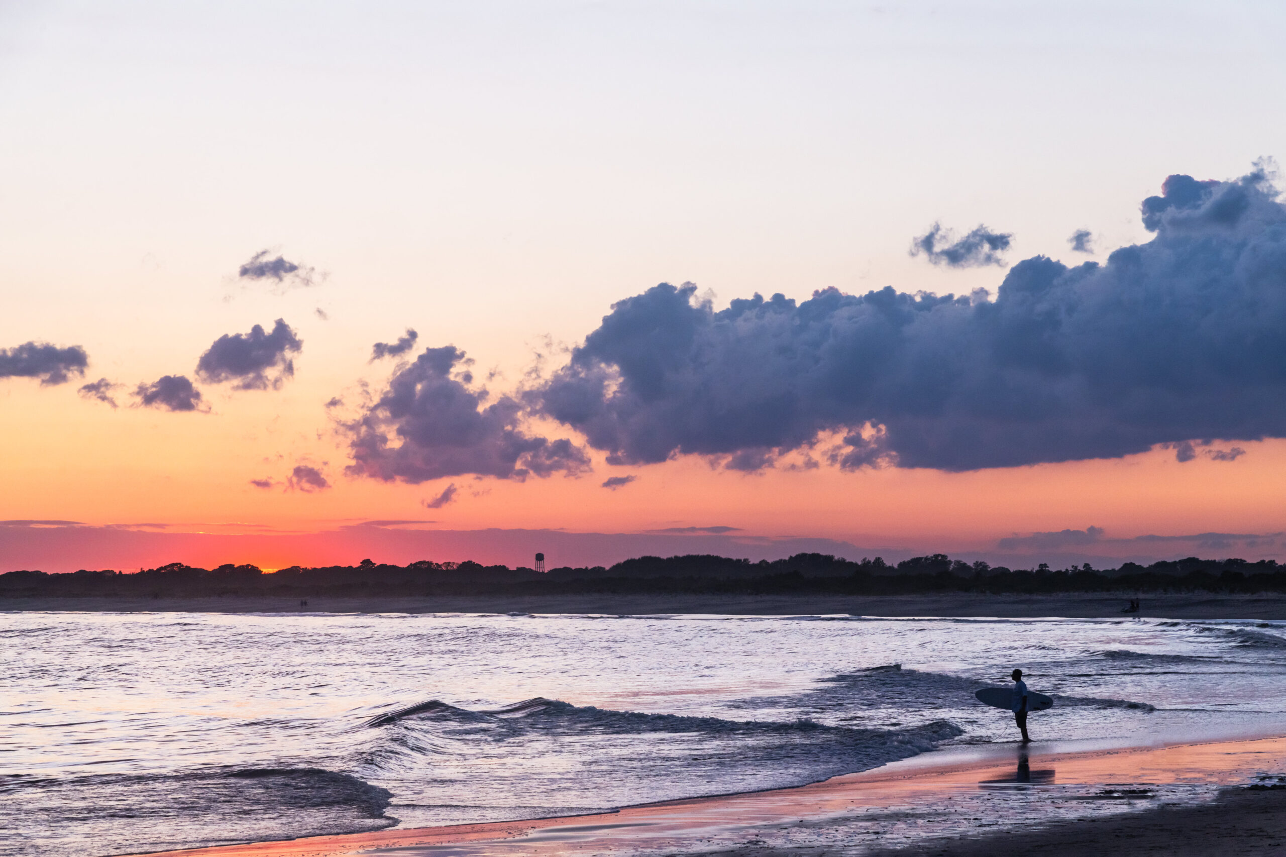 A big puffy purple cloud in a clear sky with pink at the horizon, and a surfer standing at the ocean