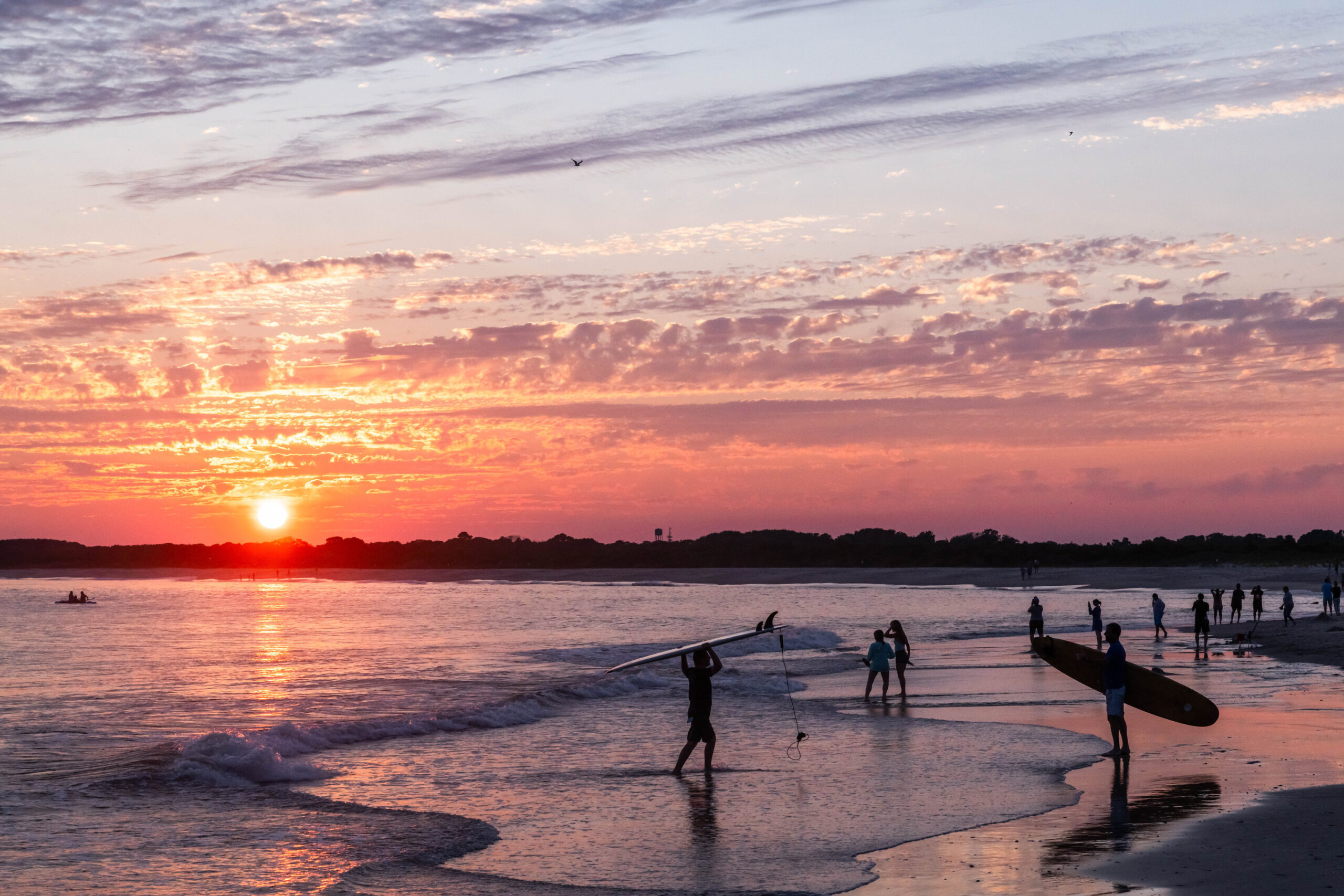 Two surfers and other people going into the ocean at sunset with a few thin clouds in the sky