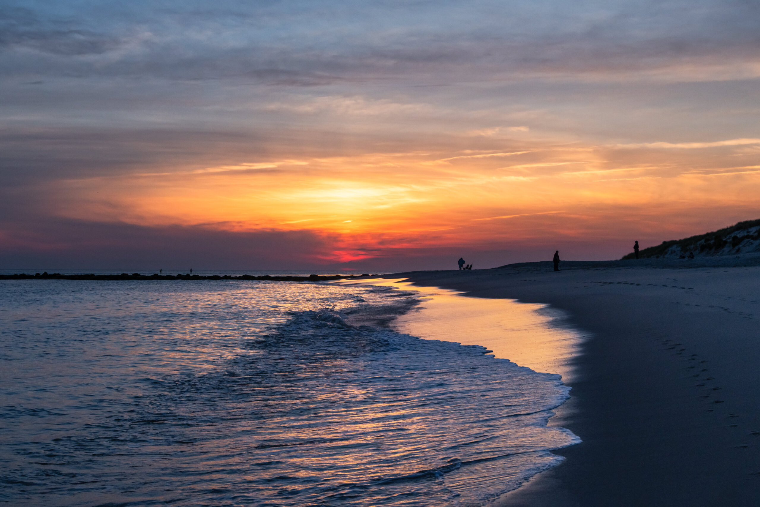 Sun shining through thin clouds in a yellow, blue, pink, and purple sky, with the ocean rushing into the shoreline at sunset